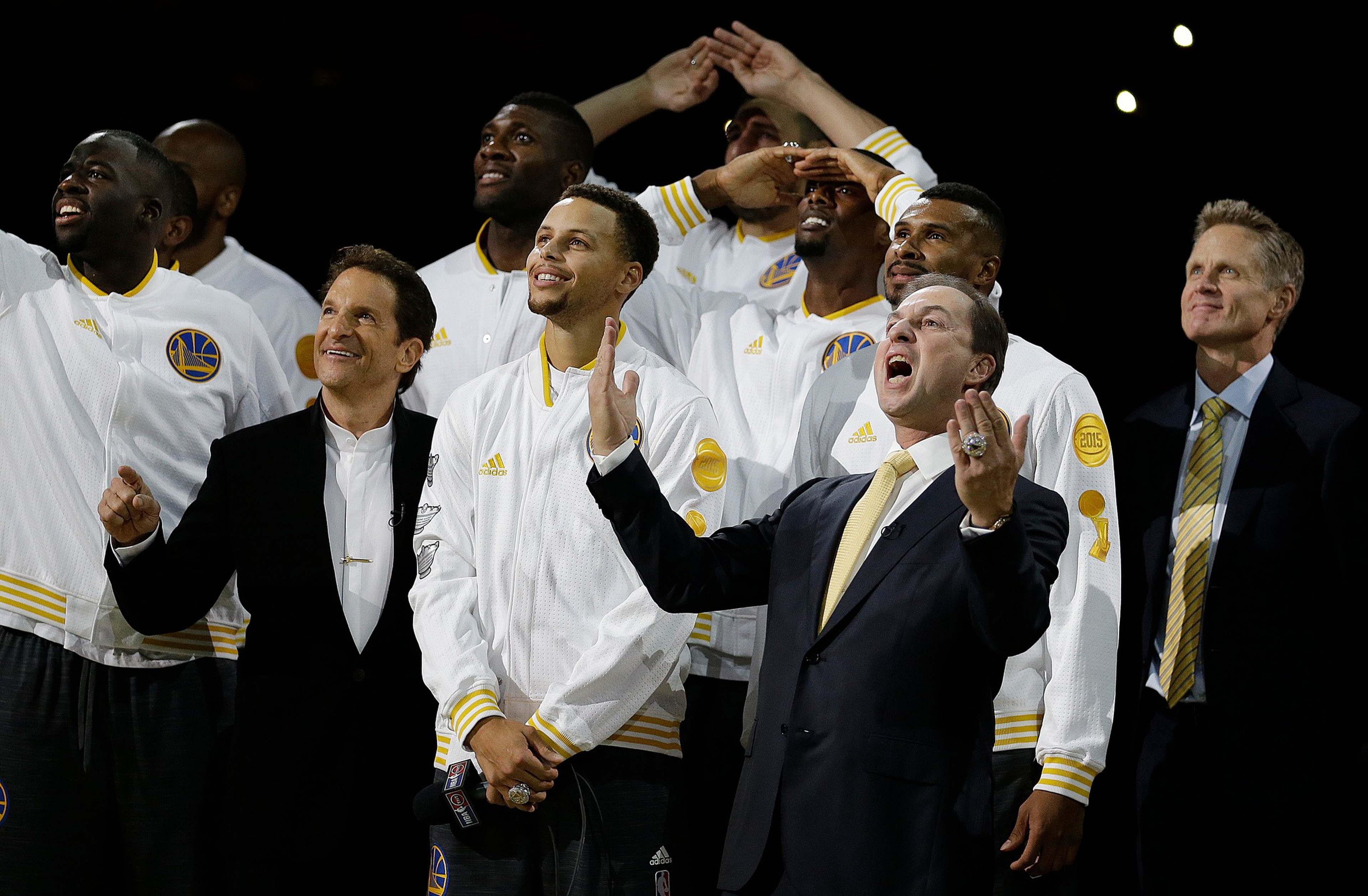 A group of Golden State Warriors players and staff in white team jackets celebrate joyfully, some pointing upward, with one man in a suit showing off a championship ring.