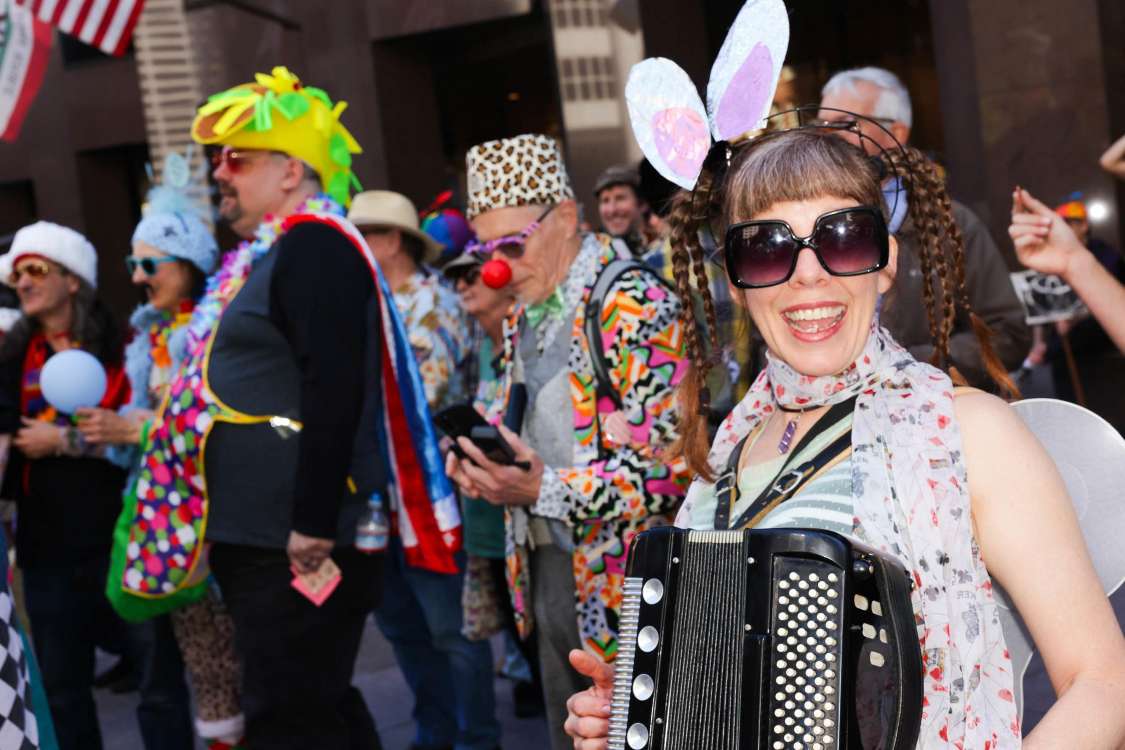 A group of joyous people in vibrant, whimsical costumes, one playing an accordion, celebrating outdoors.