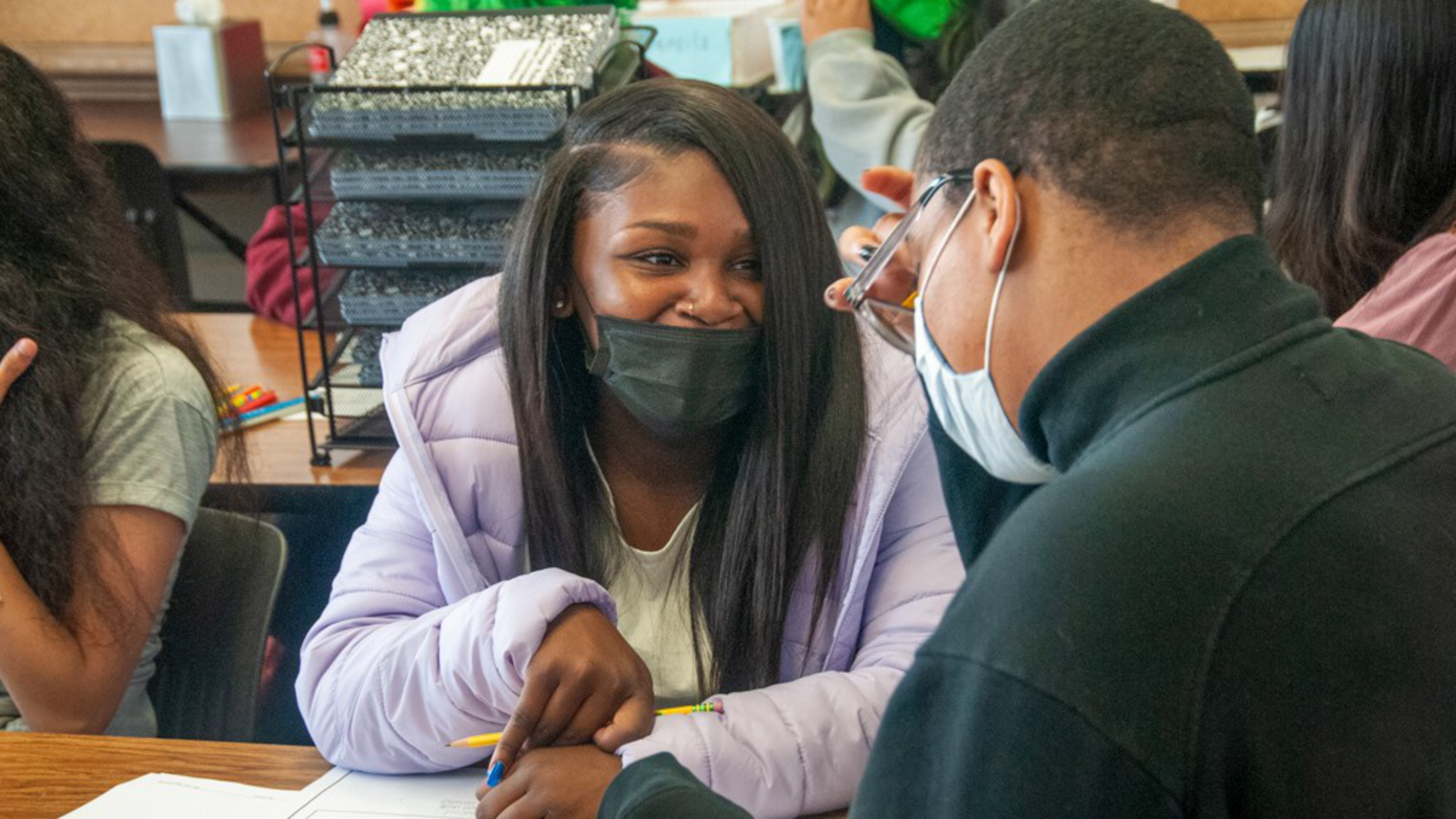 A young woman in a light purple jacket smiles at a young man wearing glasses and a face mask as they sit together at a table in a classroom.