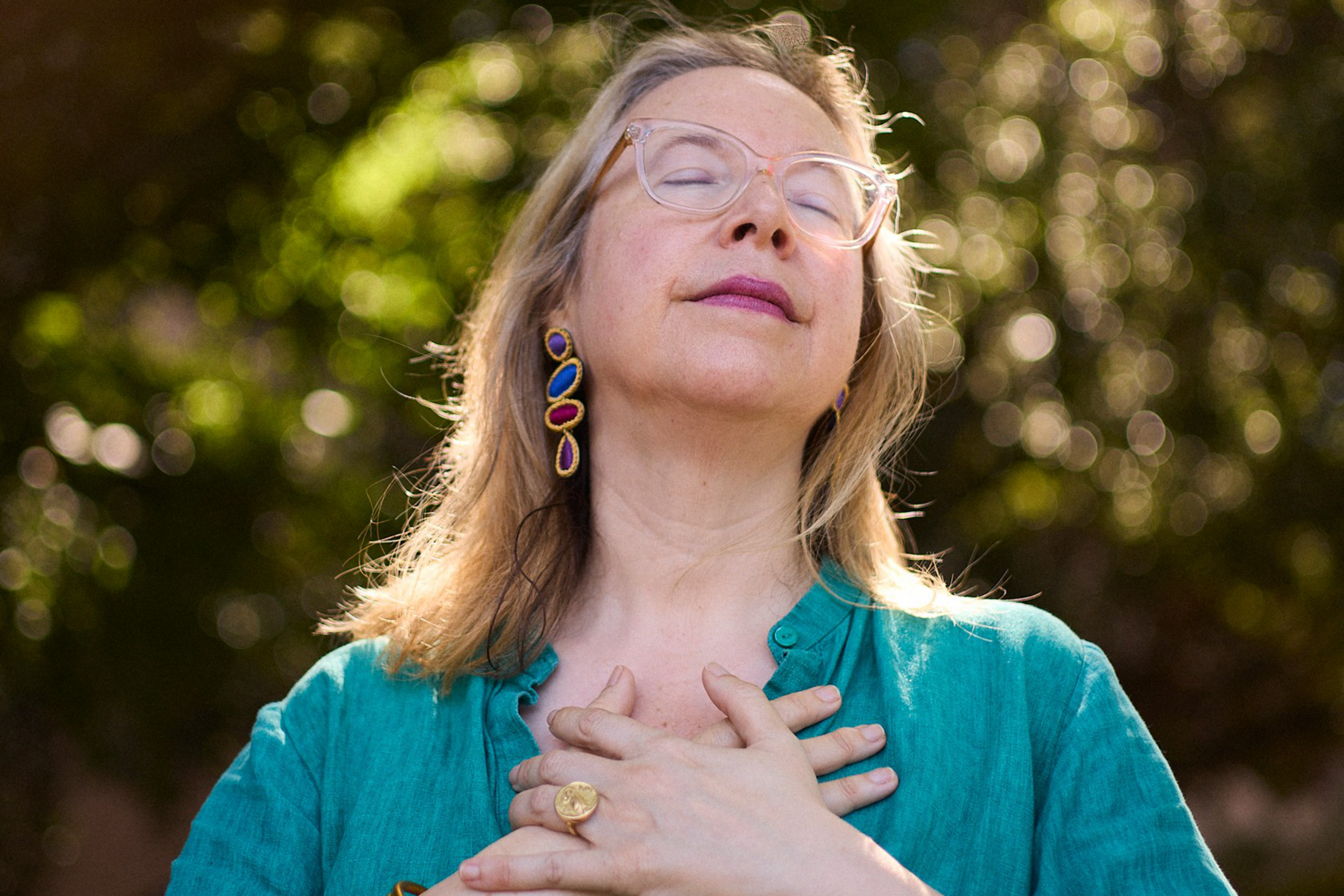 A woman with light hair and clear glasses wears a teal blouse, colorful earrings, and gold bracelets, with hands on her chest and eyes closed peacefully.