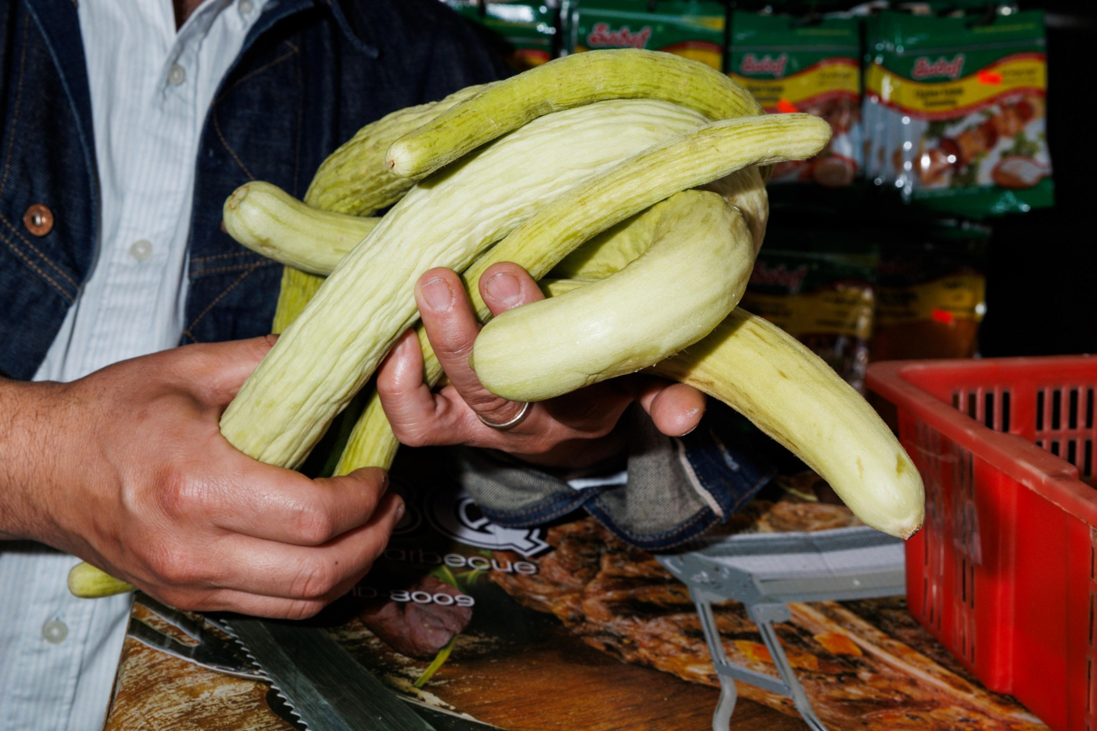 A person is holding several long, yellow-green, curved cucumbers in both hands, with packaged goods visible in the background.