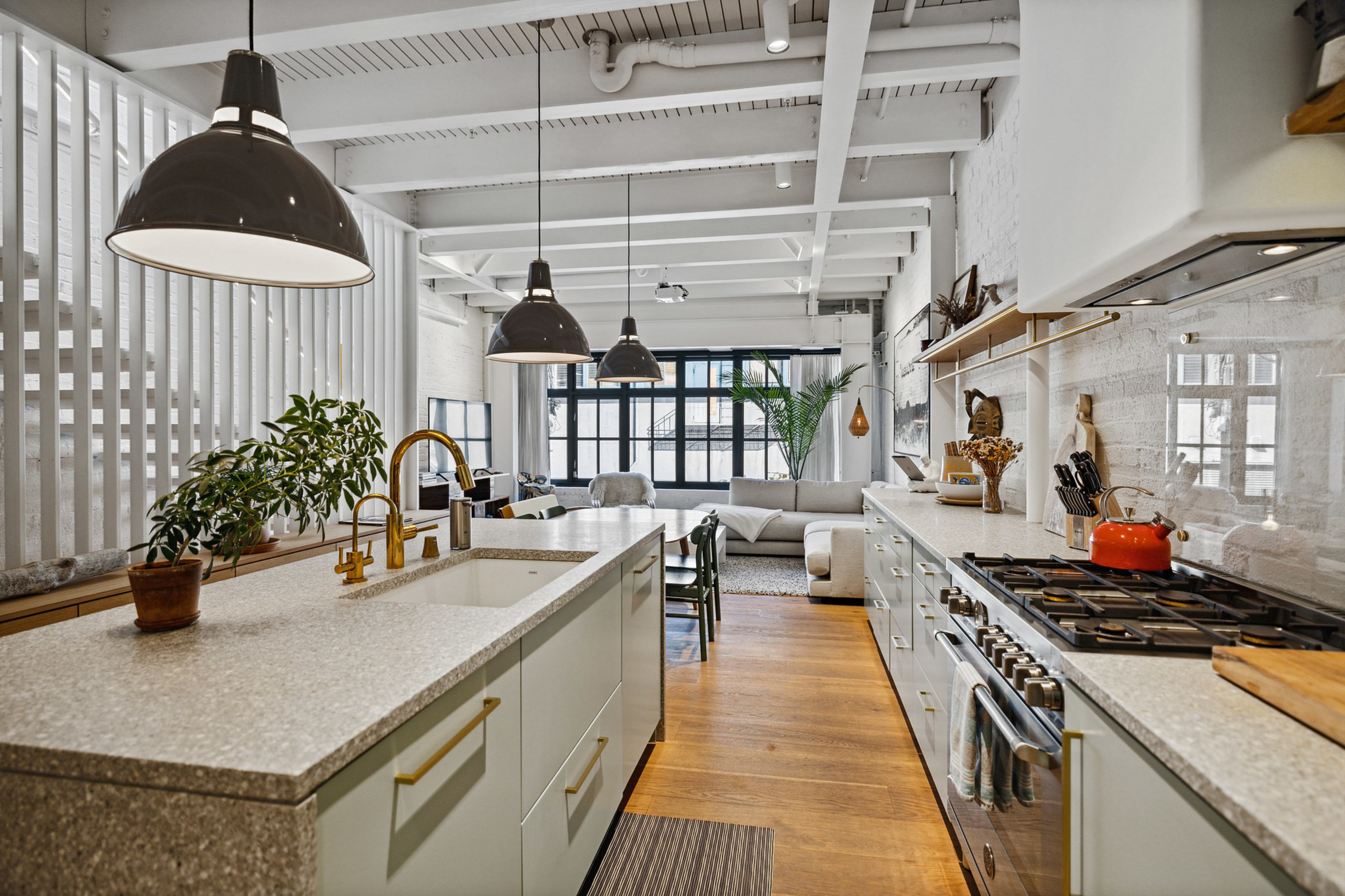 A modern kitchen with a large island, white countertops, gold fixtures, three black pendant lights, and an adjoining living area with a beige sofa.
