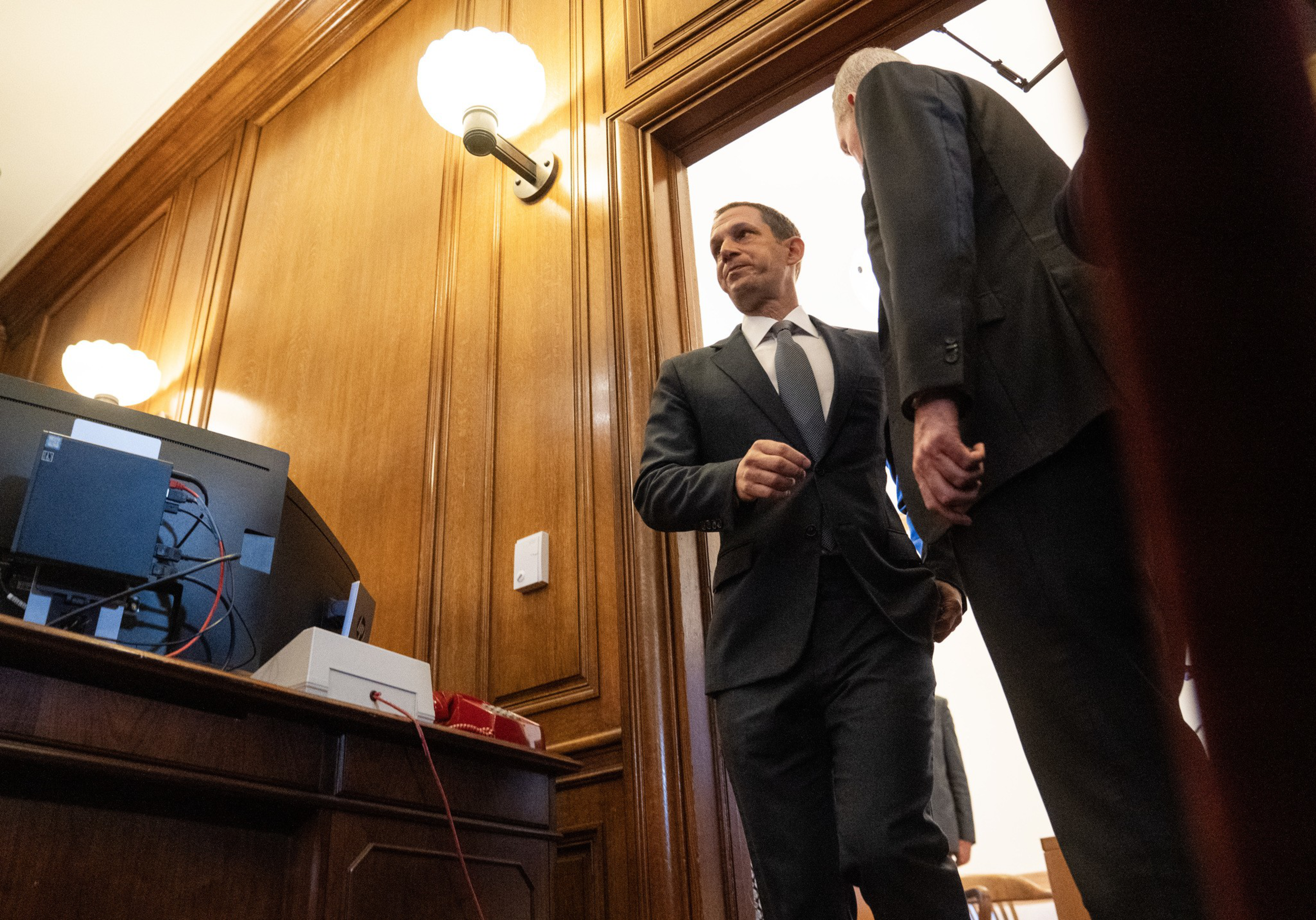 Two men in suits are standing and conversing near a wooden wall with a computer and office equipment on a desk beside them.