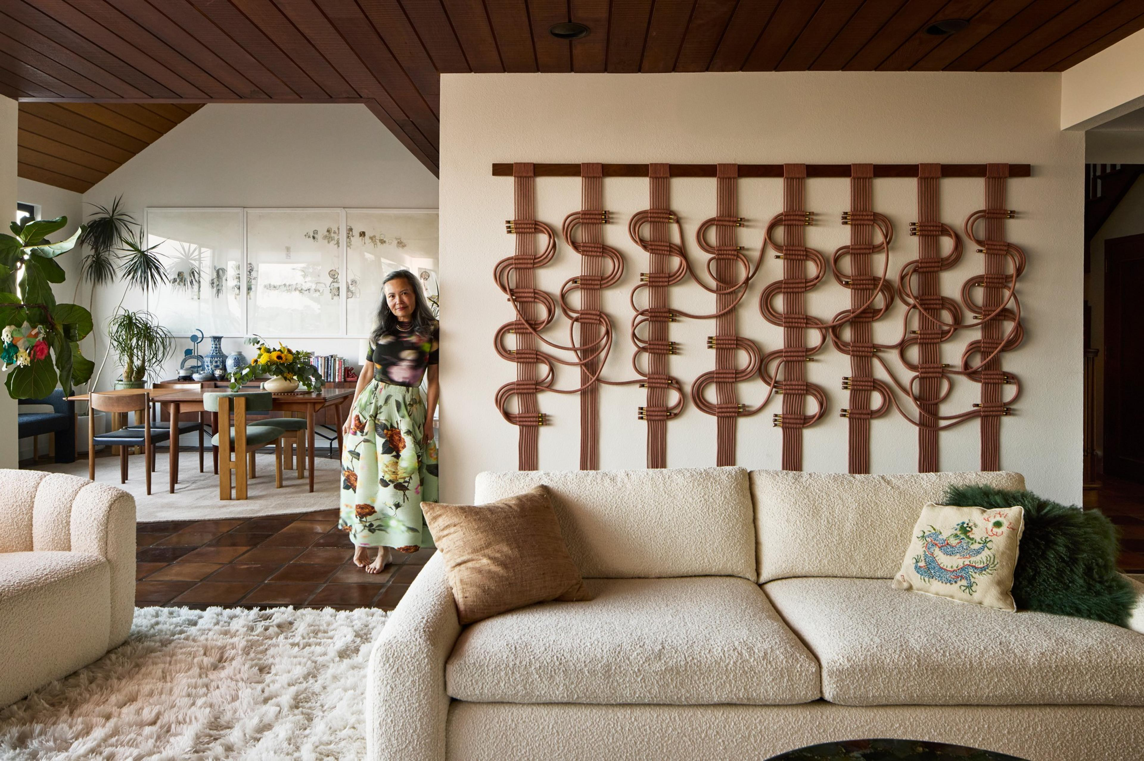 A beige sofa and armchair sit on a shaggy rug with a large, intricate wooden wall sculpture and a woman standing near a dining table with plants and artwork behind her.