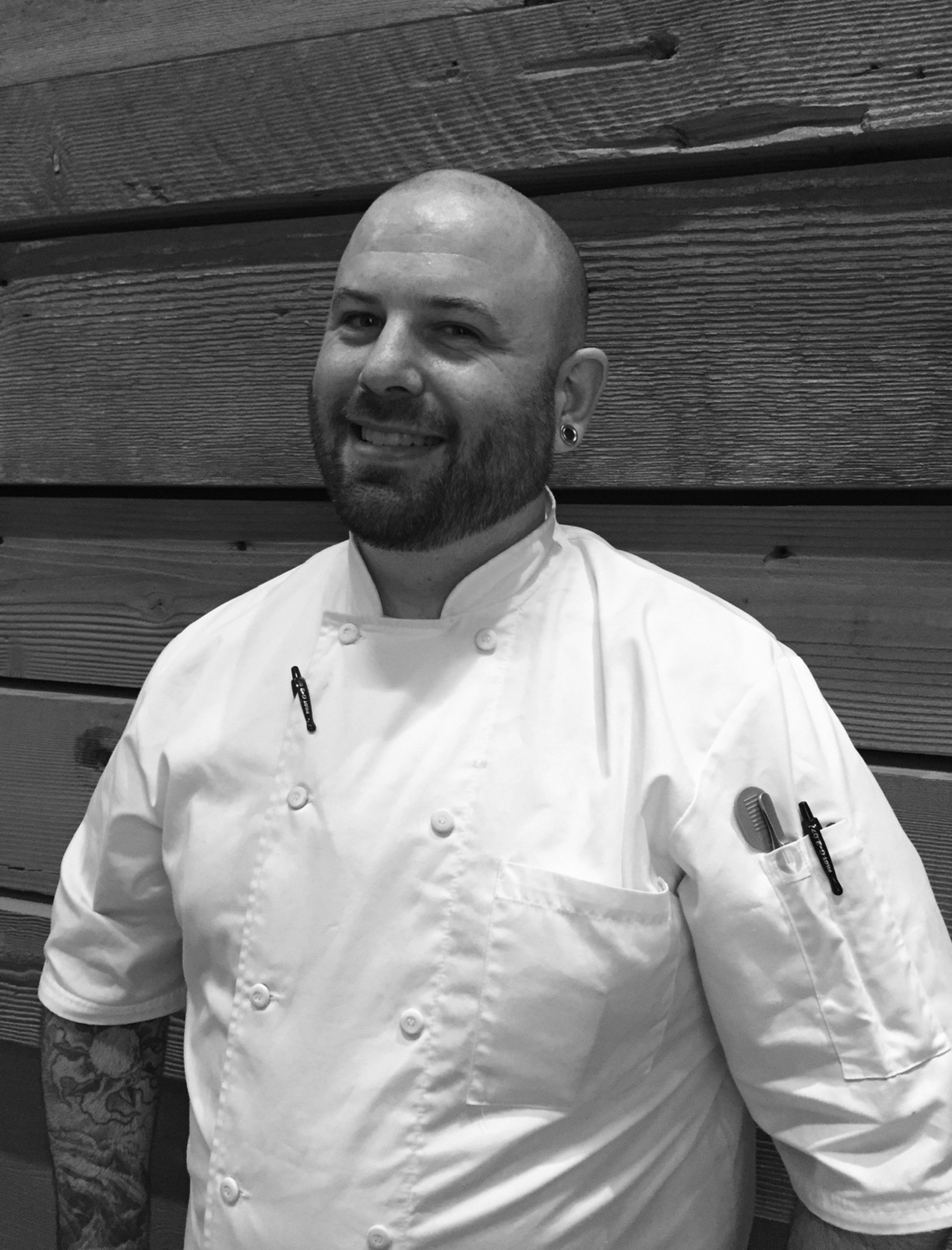 A smiling bald man with a beard wears a white chef's coat with pens and a comb in the pockets, standing in front of wooden planks.