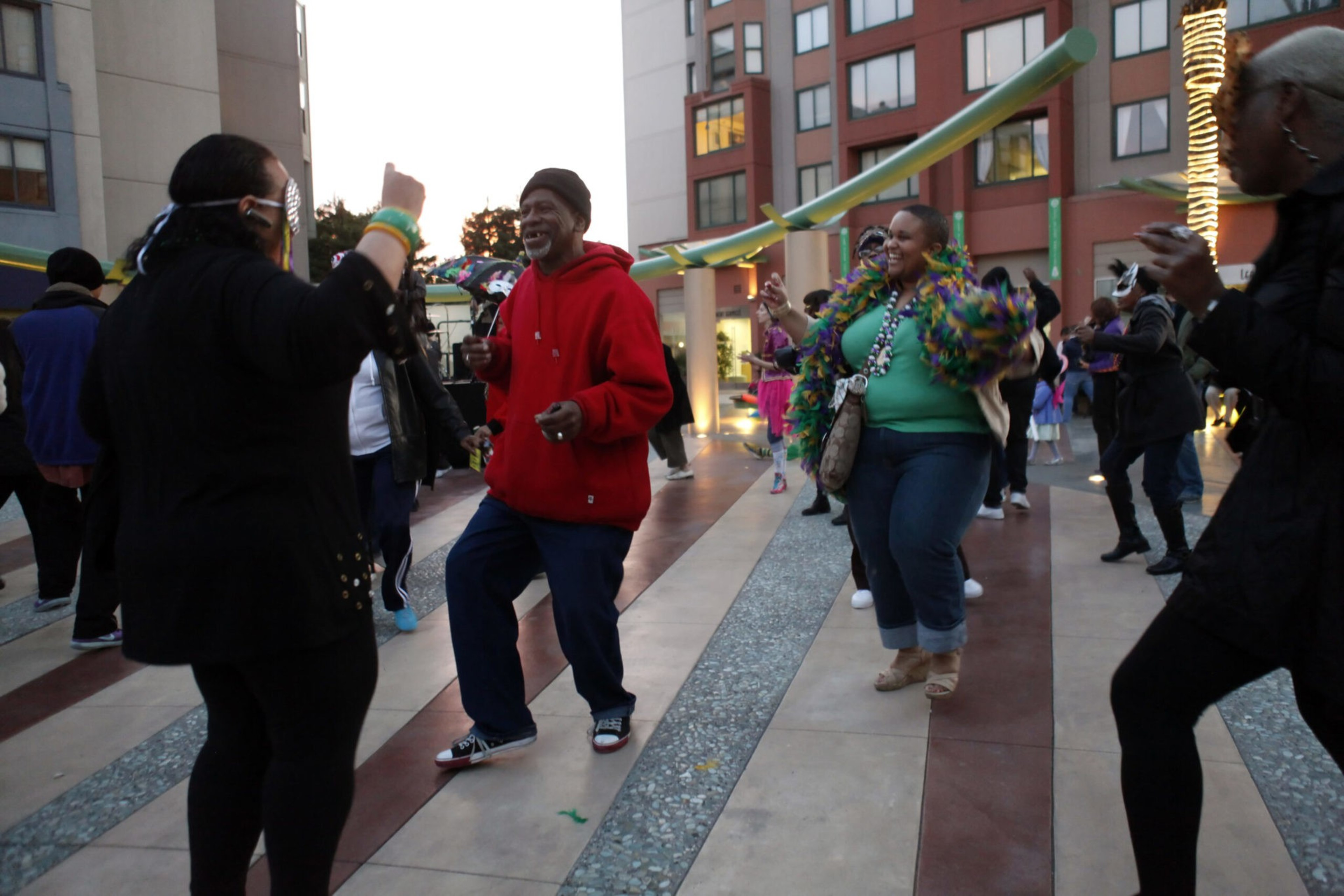 A group of people dancing outdoors on a paved area between buildings at dusk, with some wearing colorful accessories and smiling.