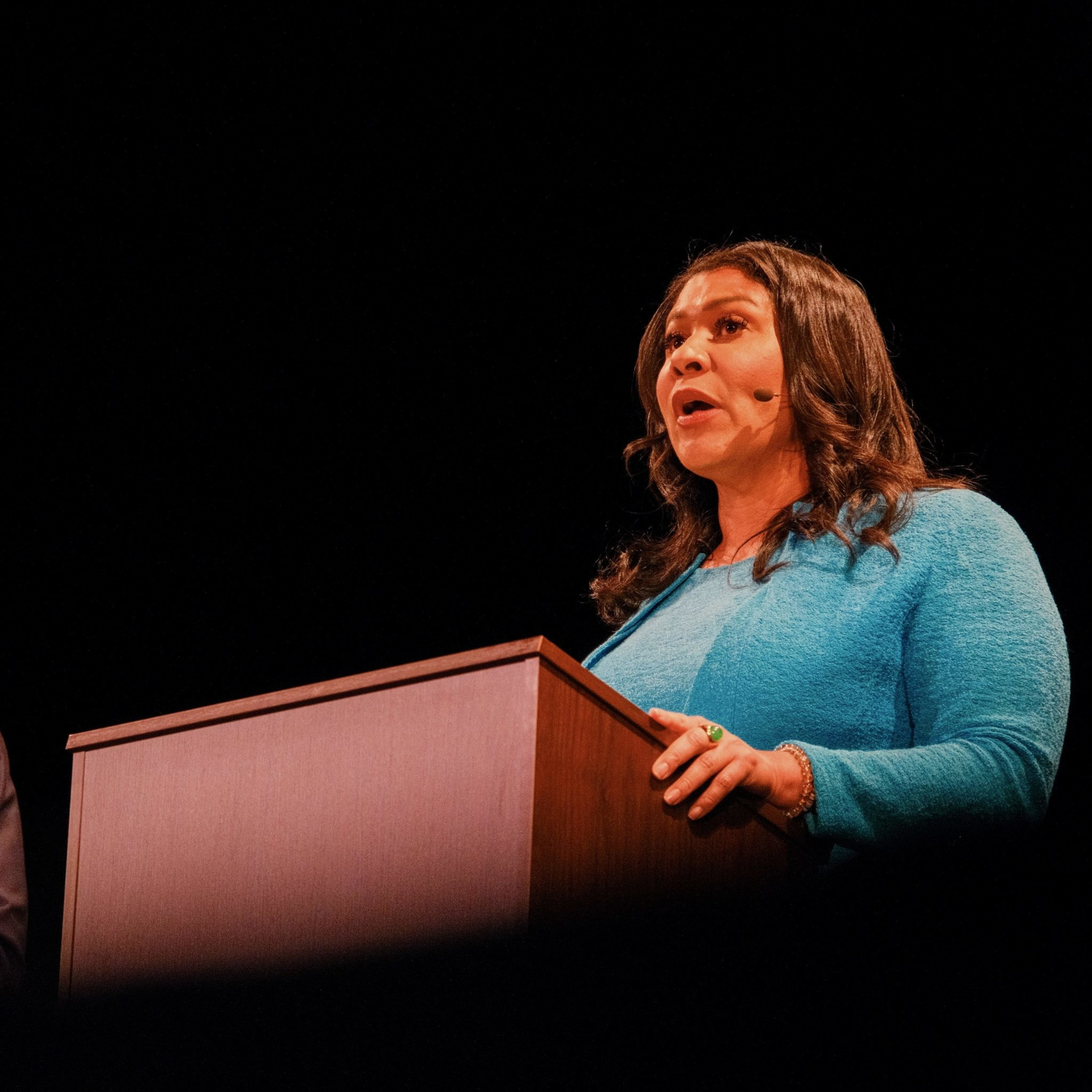 A woman is speaking at a podium, wearing a blue top, with a headset microphone on. The background is completely black, focusing attention on her.