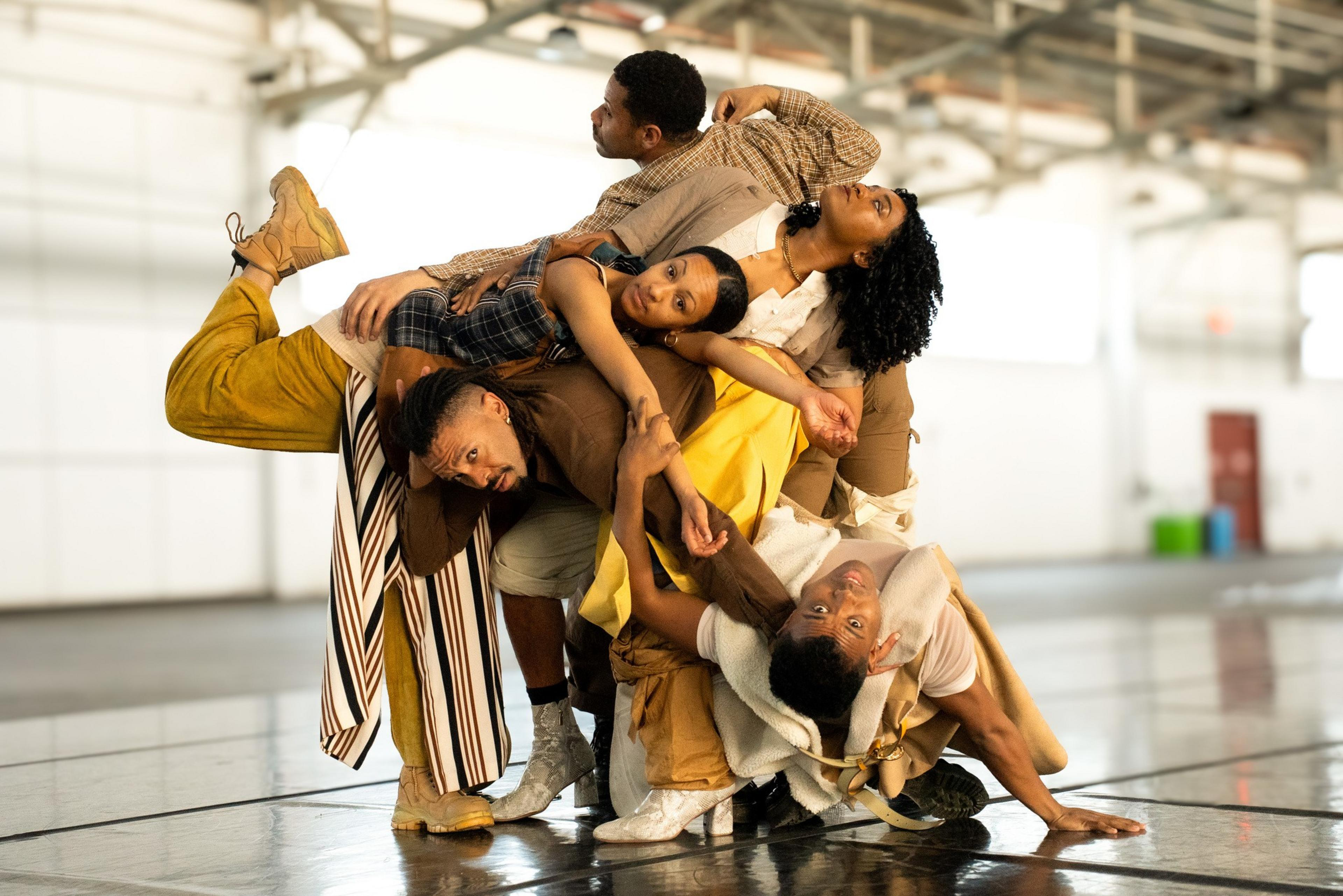 Five dancers interlock in a dynamic pose, balancing on each other with intense expressions inside a spacious, industrial-style dance studio.
