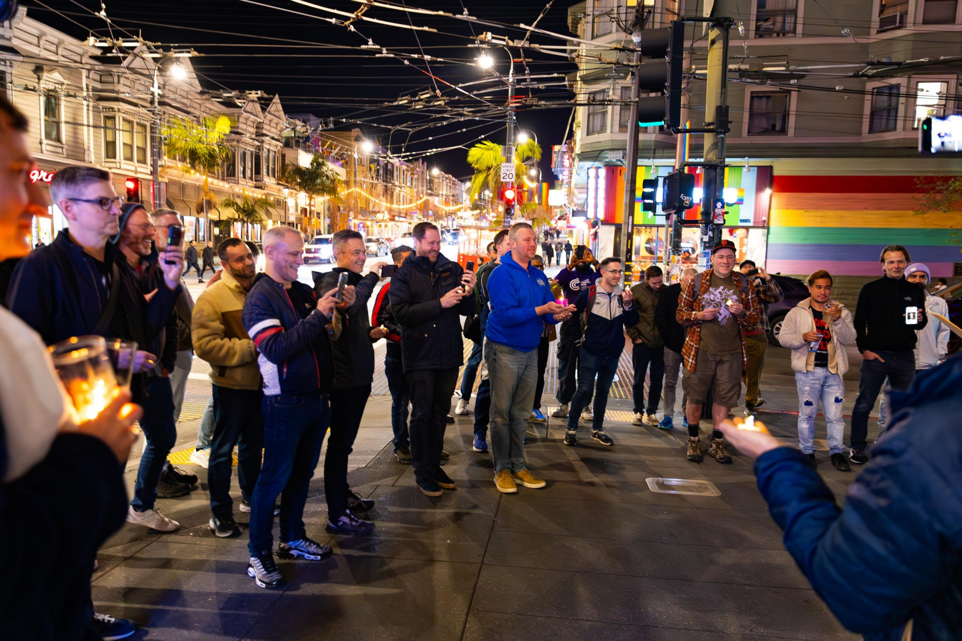 A group of people stand on a lively street at night, holding lit candles. The background features colorful lights and a building with a rainbow mural.