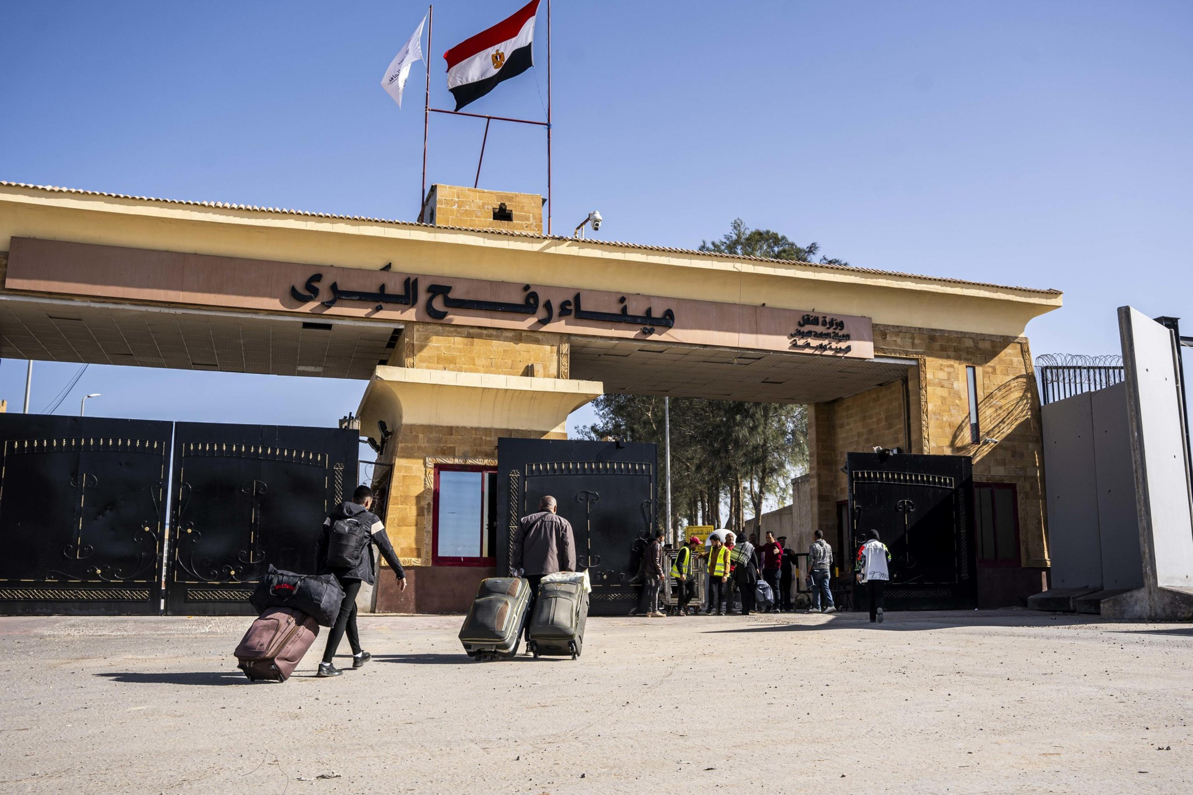 Travelers with luggage walk towards a gate beneath a building with flags and Arab script signage.