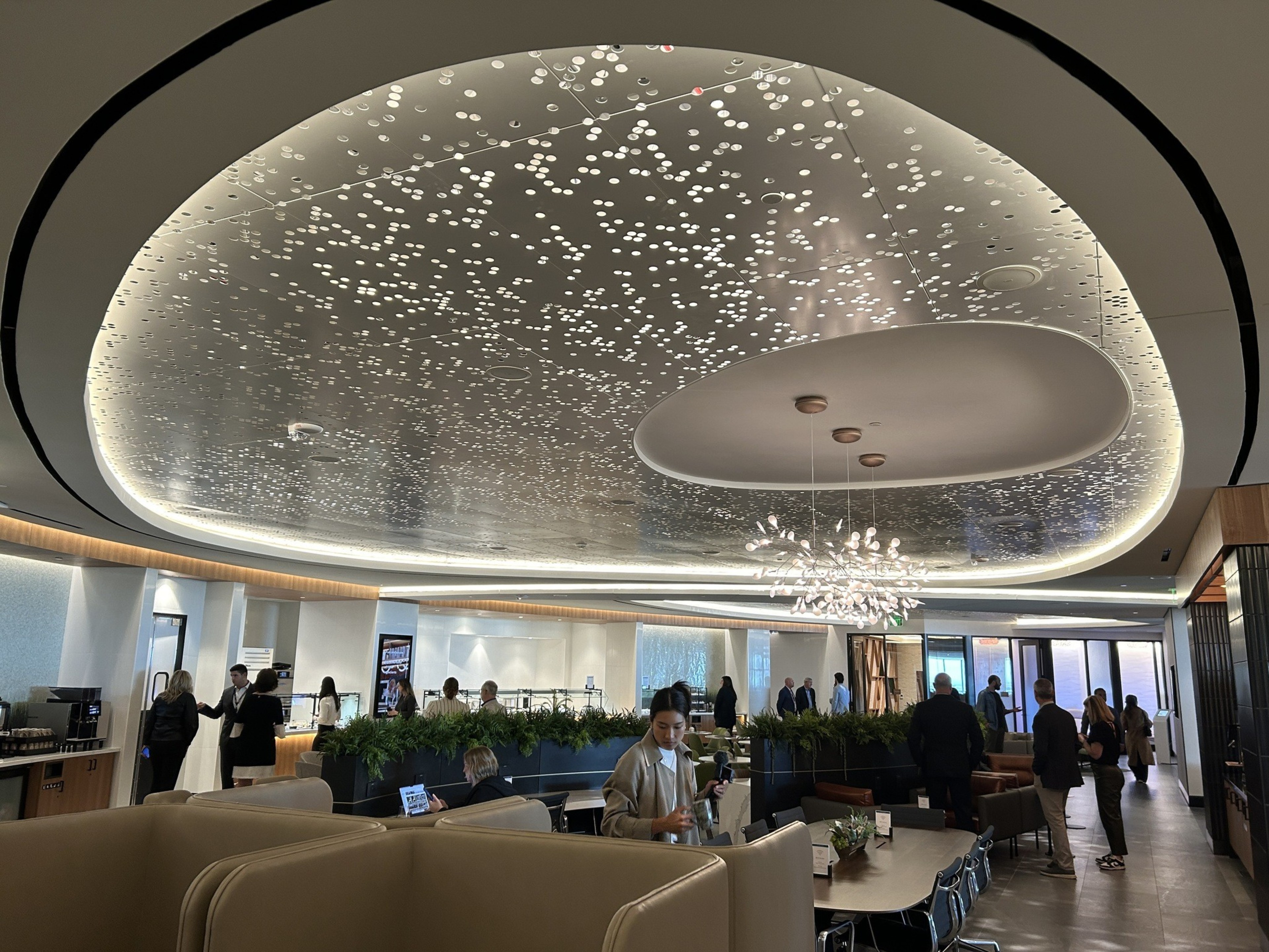 The image shows a modern, well-lit lounge with a decorative ceiling featuring small, illuminated dots and a unique chandelier. People are socializing and working at tables.