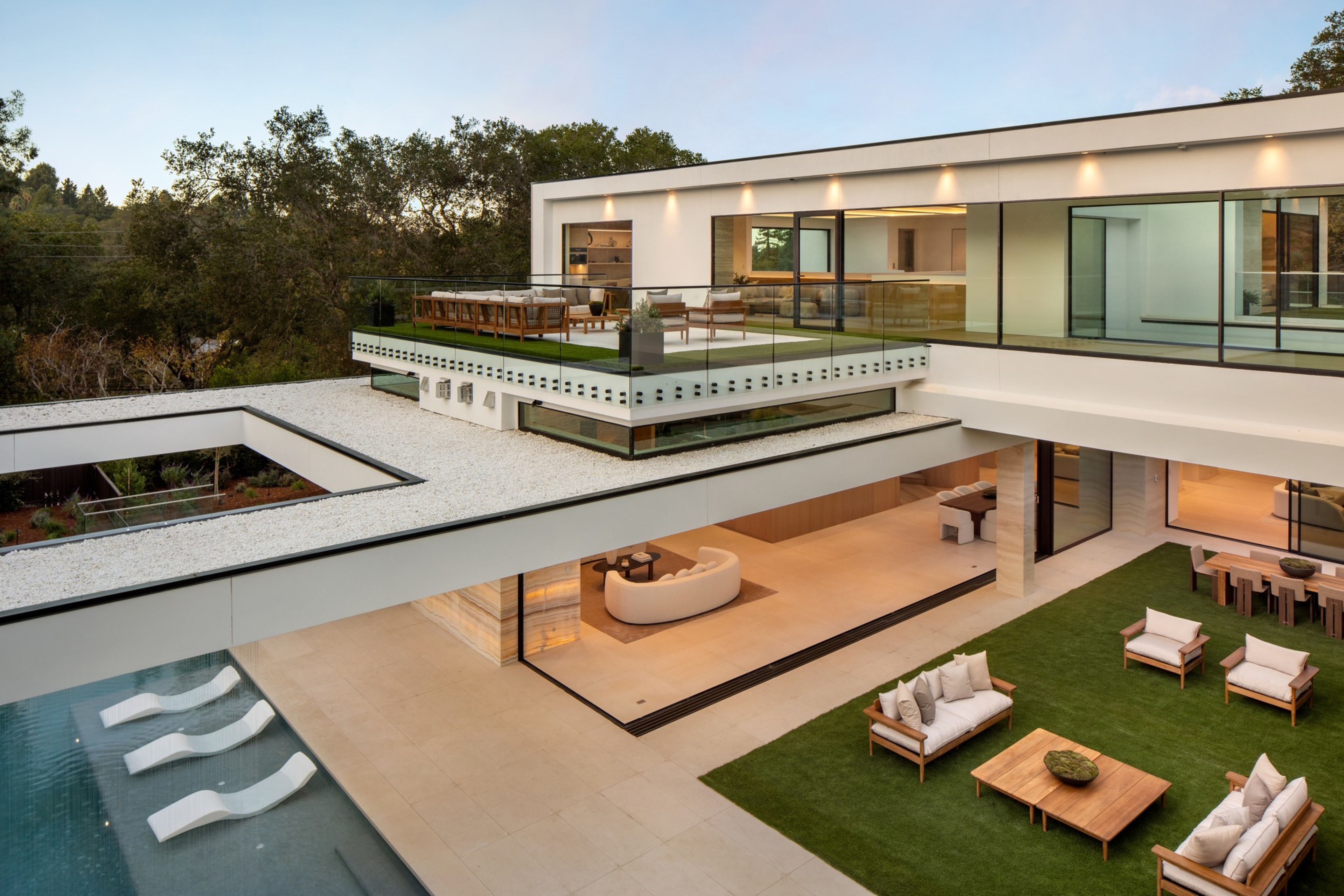 A modern two-story house features a large outdoor patio with seating, a grassy area with couches, a dining table, and a pool with submerged lounge chairs.
