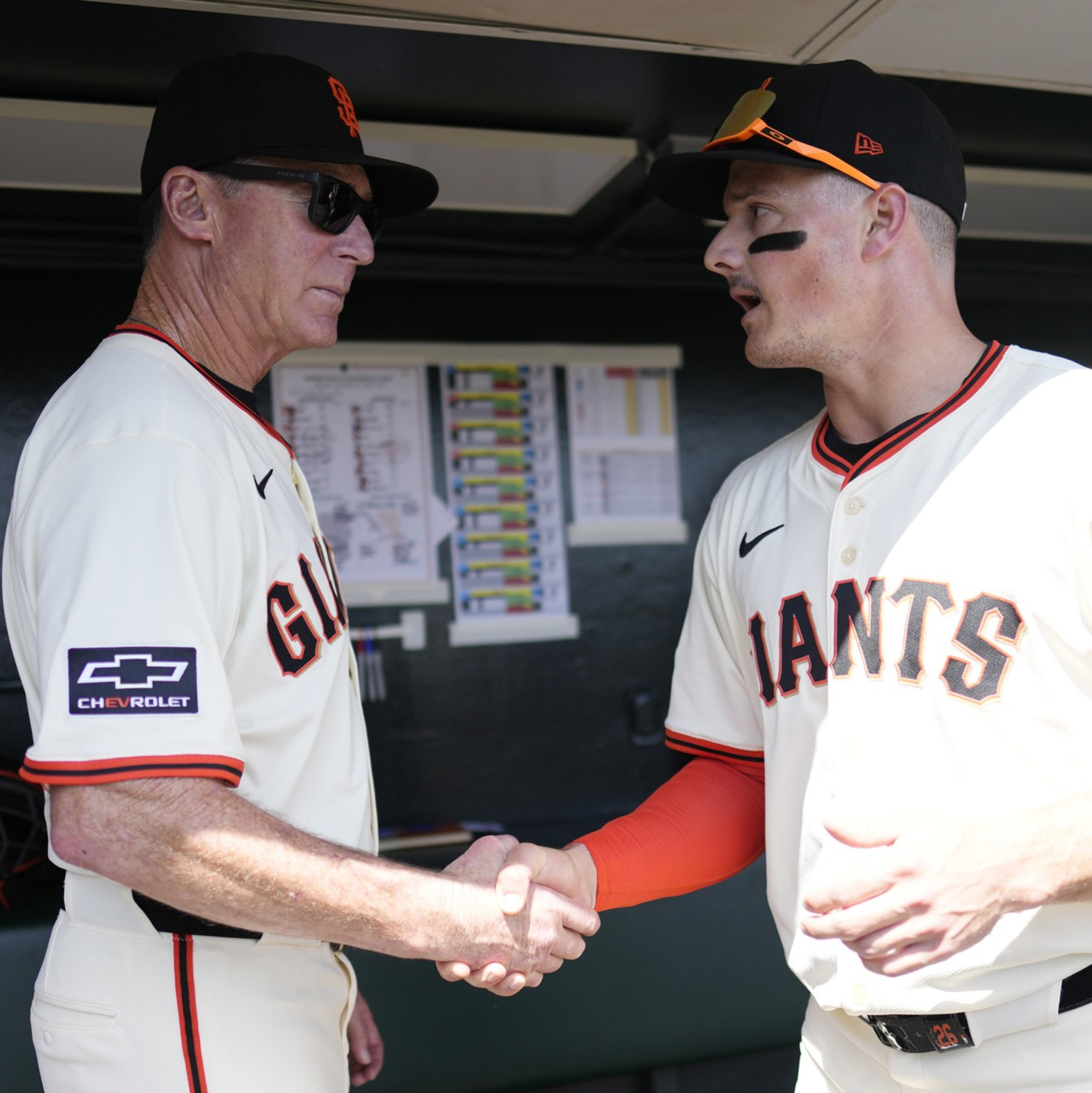 A baseball player and a coach in Giants uniforms shake hands in a dugout, with another player standing in the background.