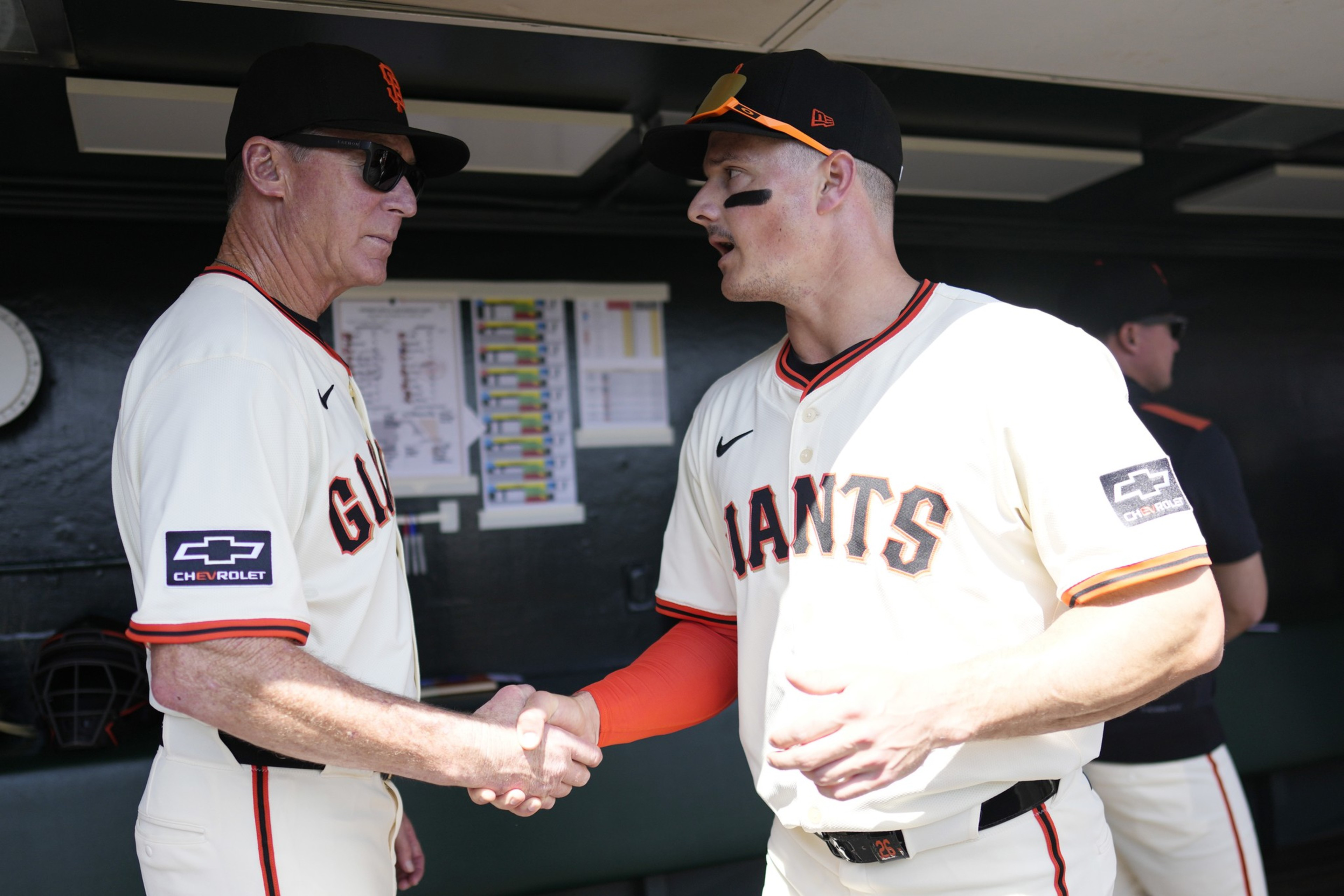 A baseball player and a coach in Giants uniforms shake hands in a dugout, with another player standing in the background.