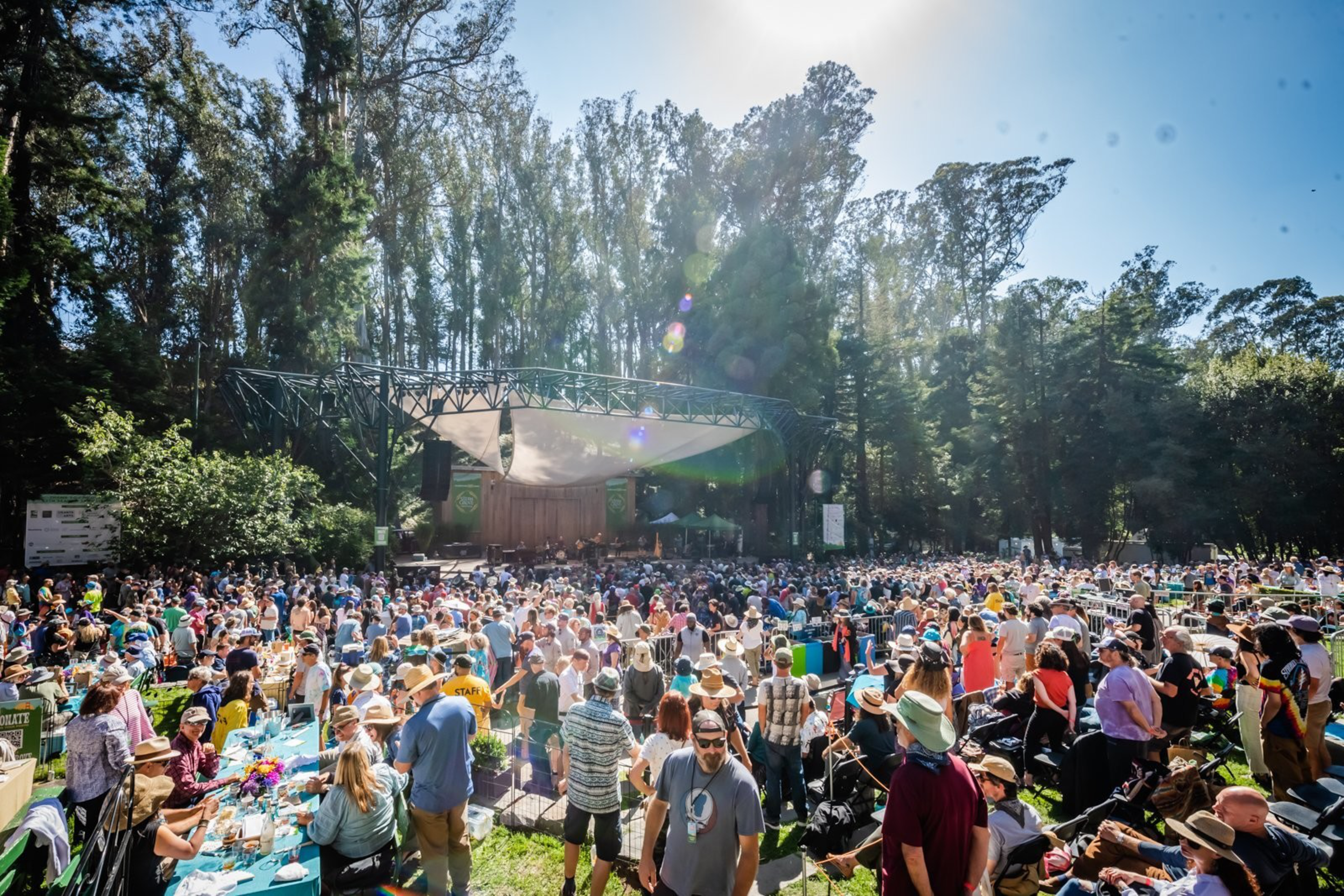 A large crowd gathers at an outdoor concert in a forested area. People are seated and standing, with many wearing hats under a sunny blue sky.