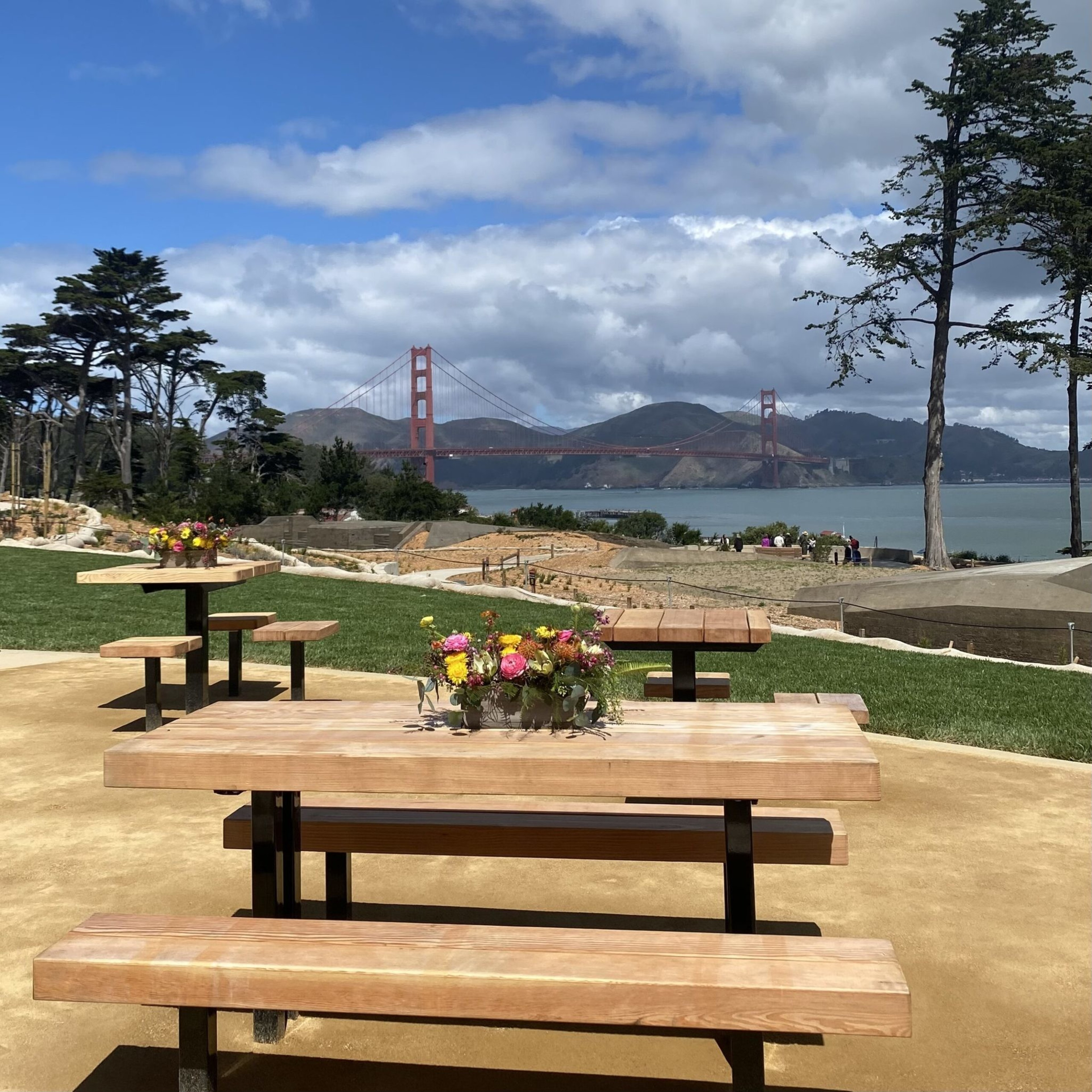 Wooden picnic tables with flower arrangements sit near grass, overlooking water and the Golden Gate Bridge under a partly cloudy sky.