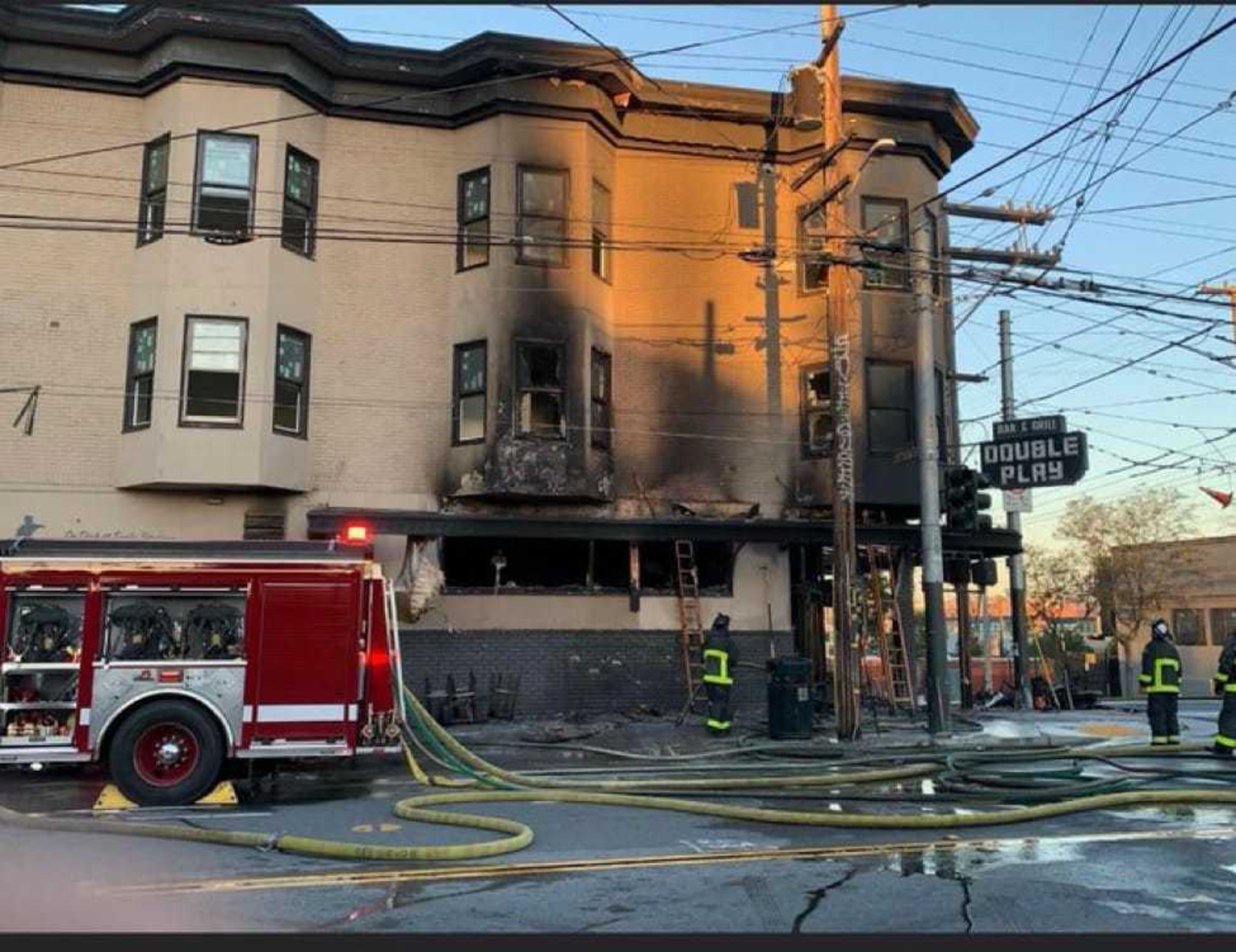 A building with severe fire damage and blackened walls has firefighters and hoses outside, alongside a fire truck and a sign reading "Double Play."