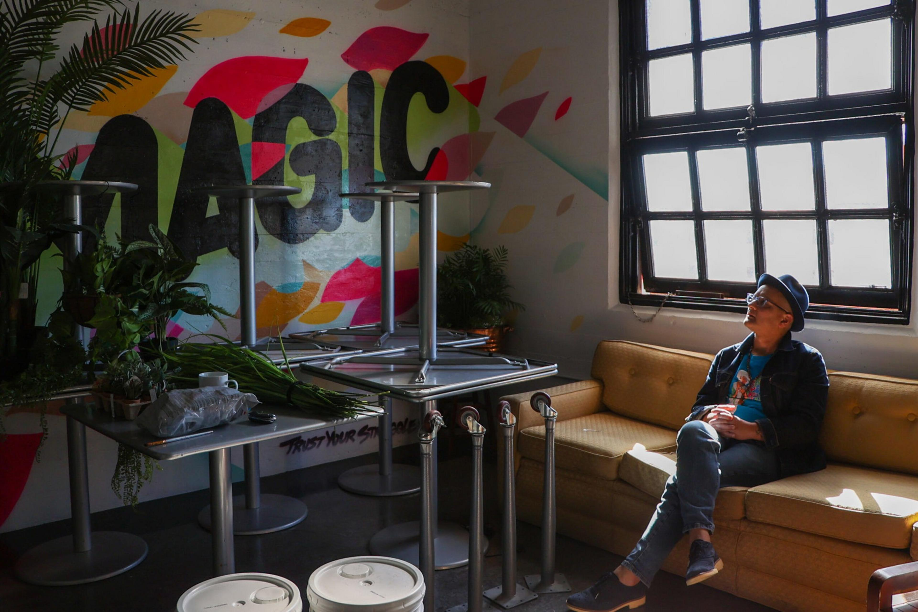 A person wearing a hat and sunglasses sits on a yellow couch, looking out a large window with light streaming in, near tables and plants with "MAGIC" graffiti.