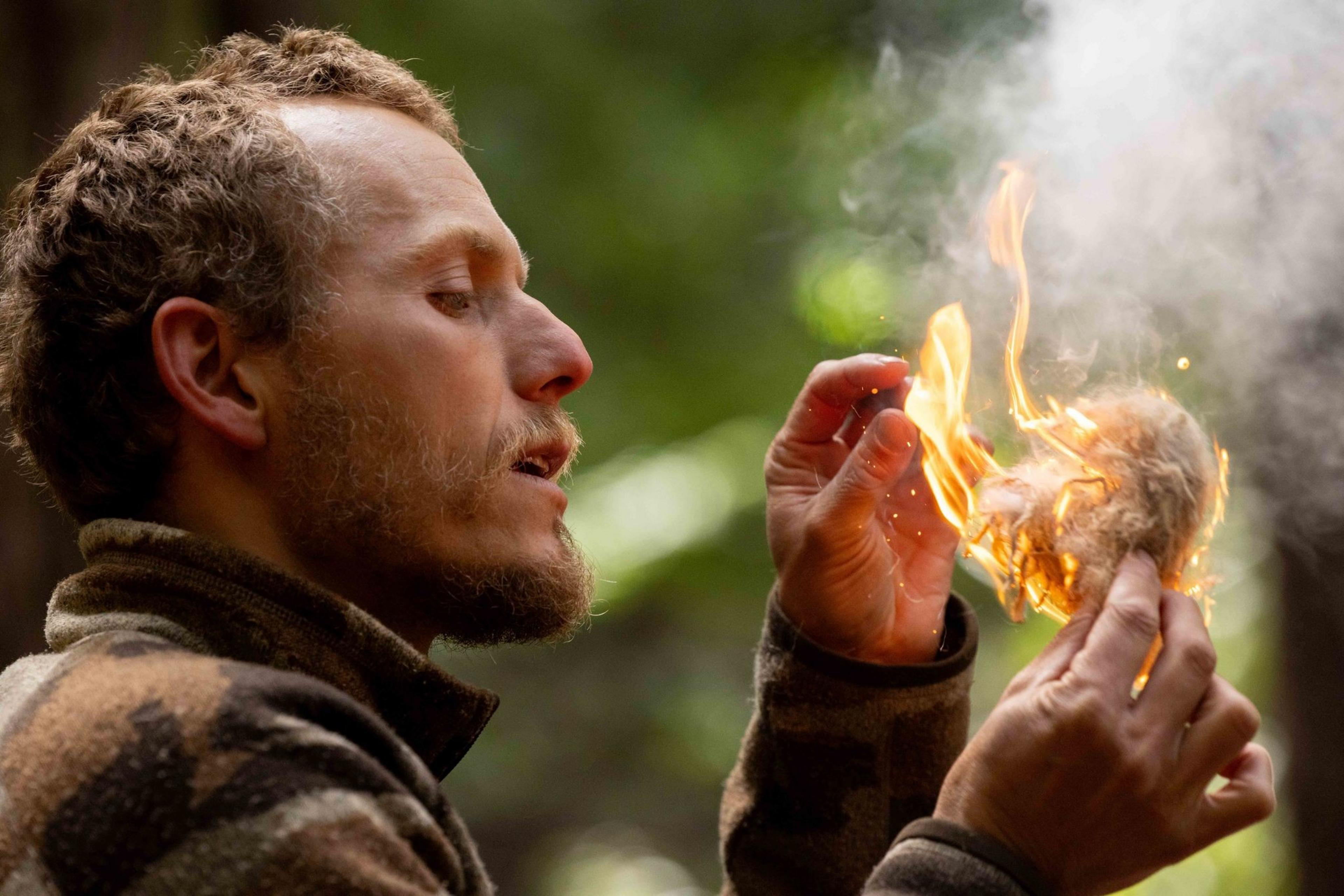 A man with short hair and a beard is carefully holding a small flame with both hands, seemingly igniting some kind of material, in an outdoor setting.