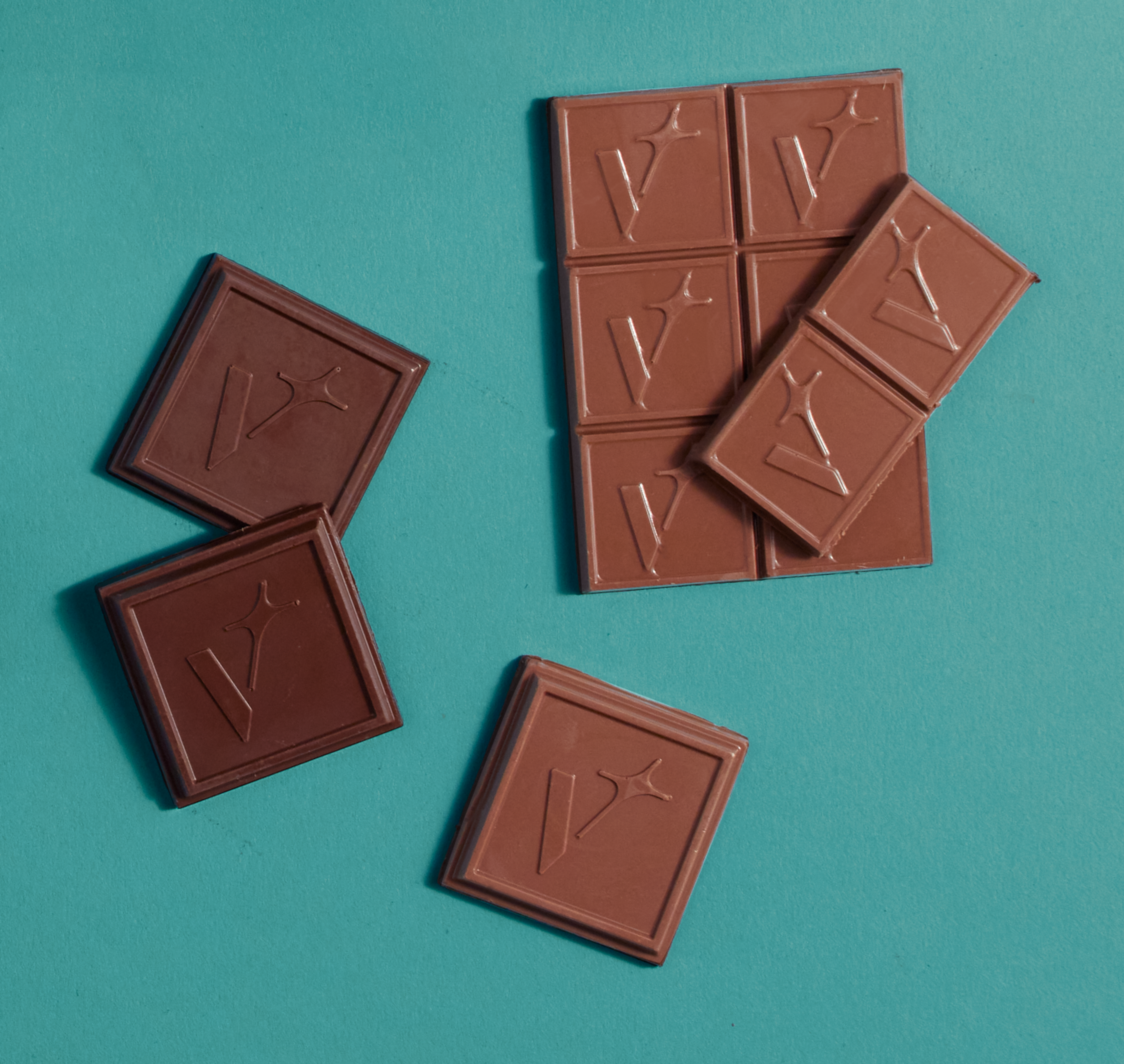 Six milk chocolate squares and three dark chocolate squares are placed on a teal surface, each embossed with a stylized "V" and a star.