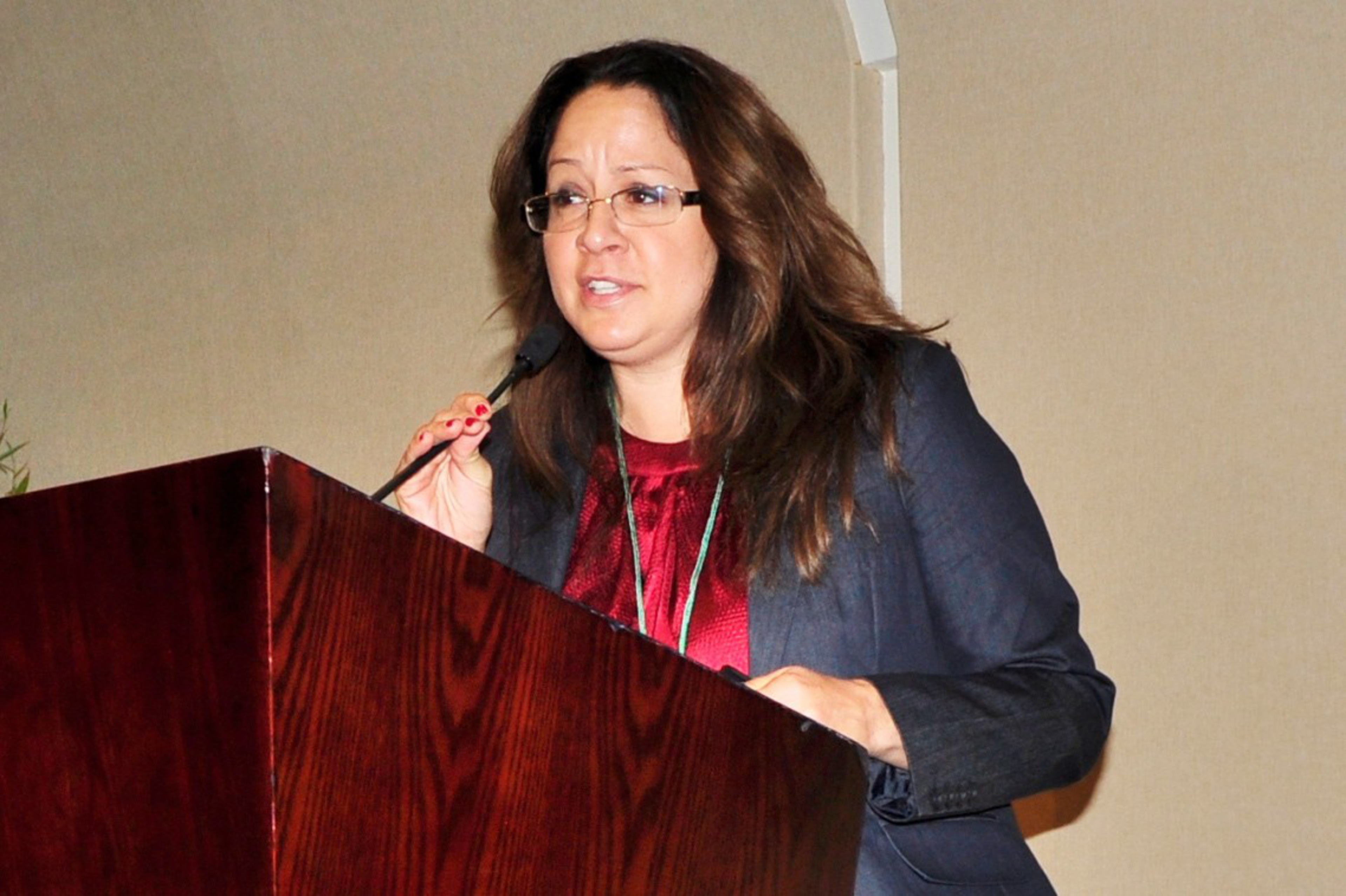 A person in a red shirt and blue coat talks at the podium.