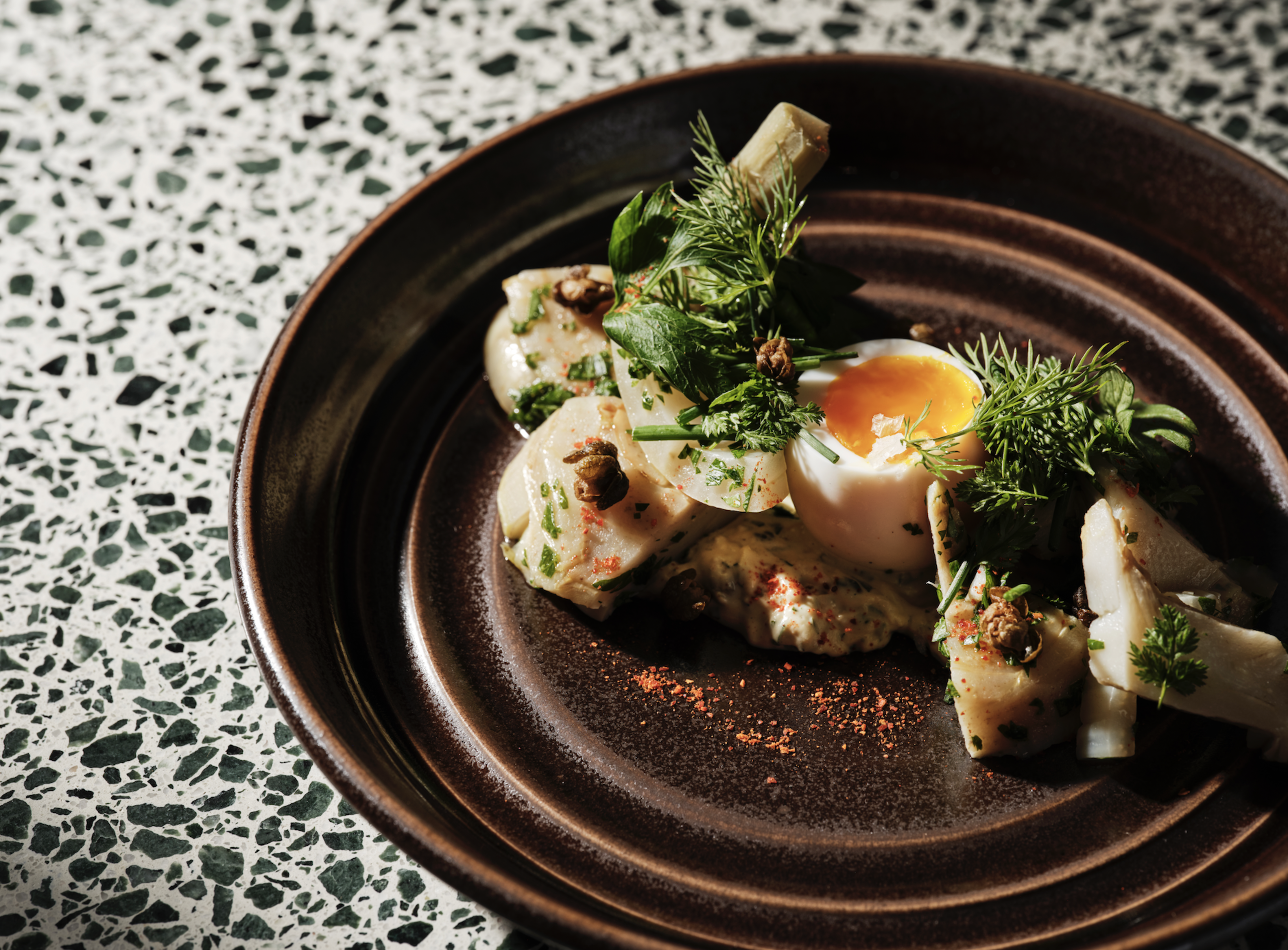 A soft-boiled egg with a runny yolk is surrounded by seasoned vegetables, fresh herbs, and a creamy sauce on a dark, round plate.