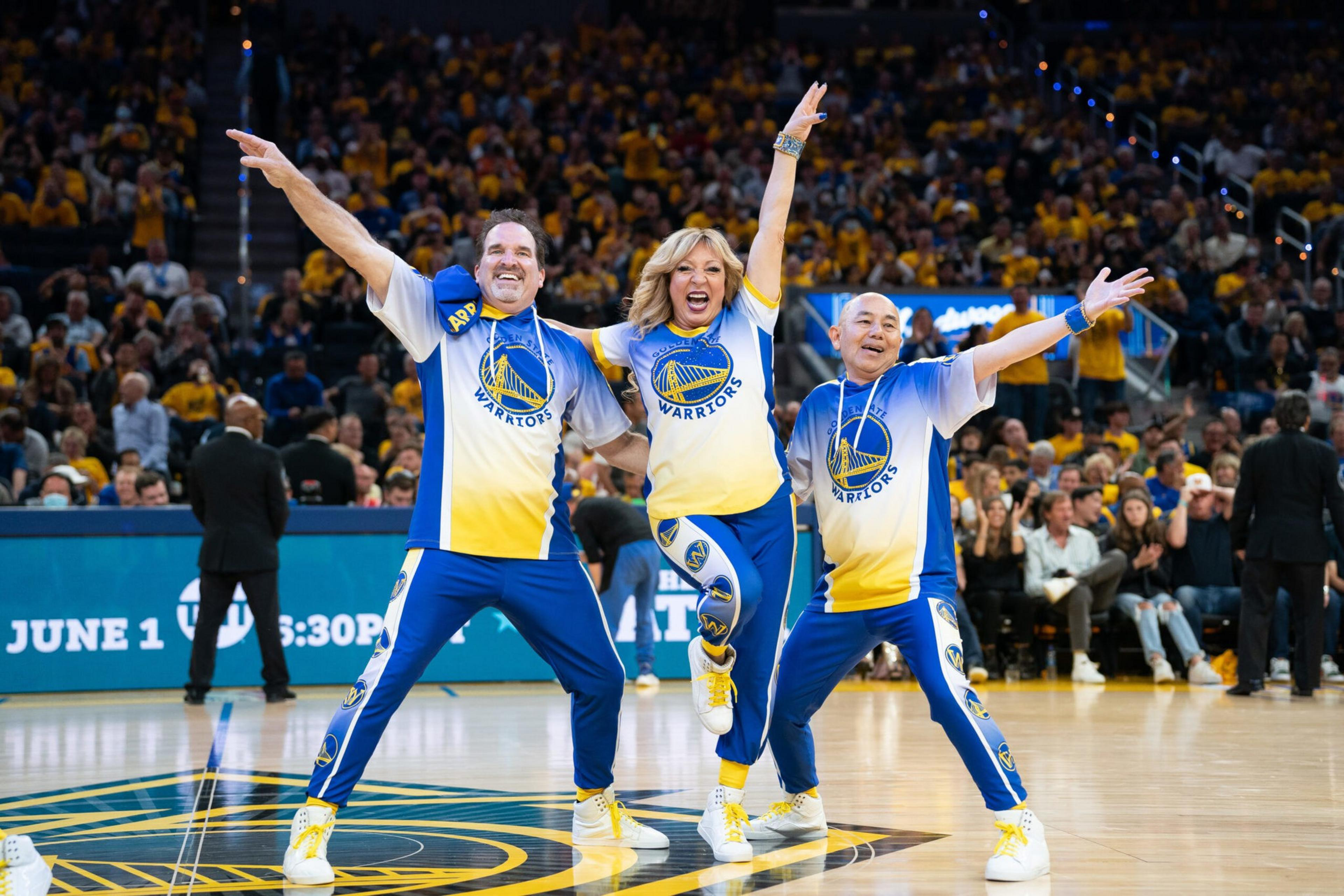 Three people in Golden State Warriors outfits strike energetic dance poses on a basketball court with a cheering crowd in the background.