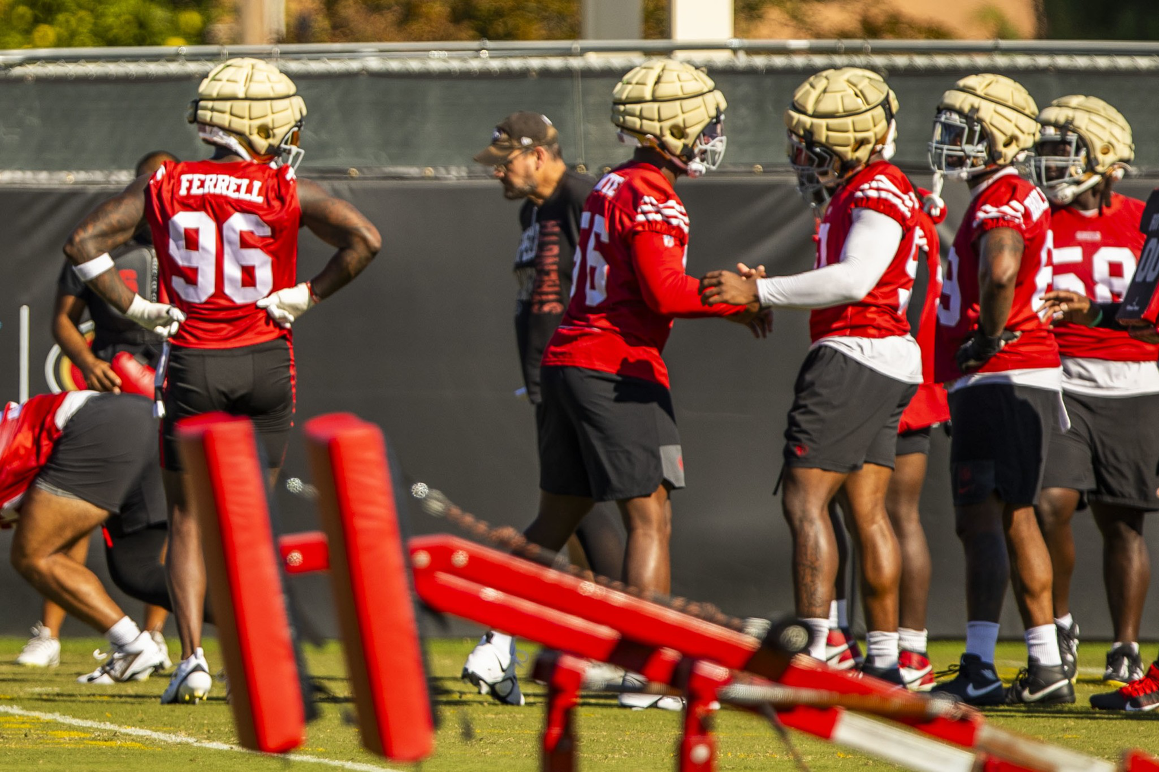 Football players in red practice jerseys and padded helmets stand on a field, preparing for drills with a coach nearby and training equipment in the foreground.