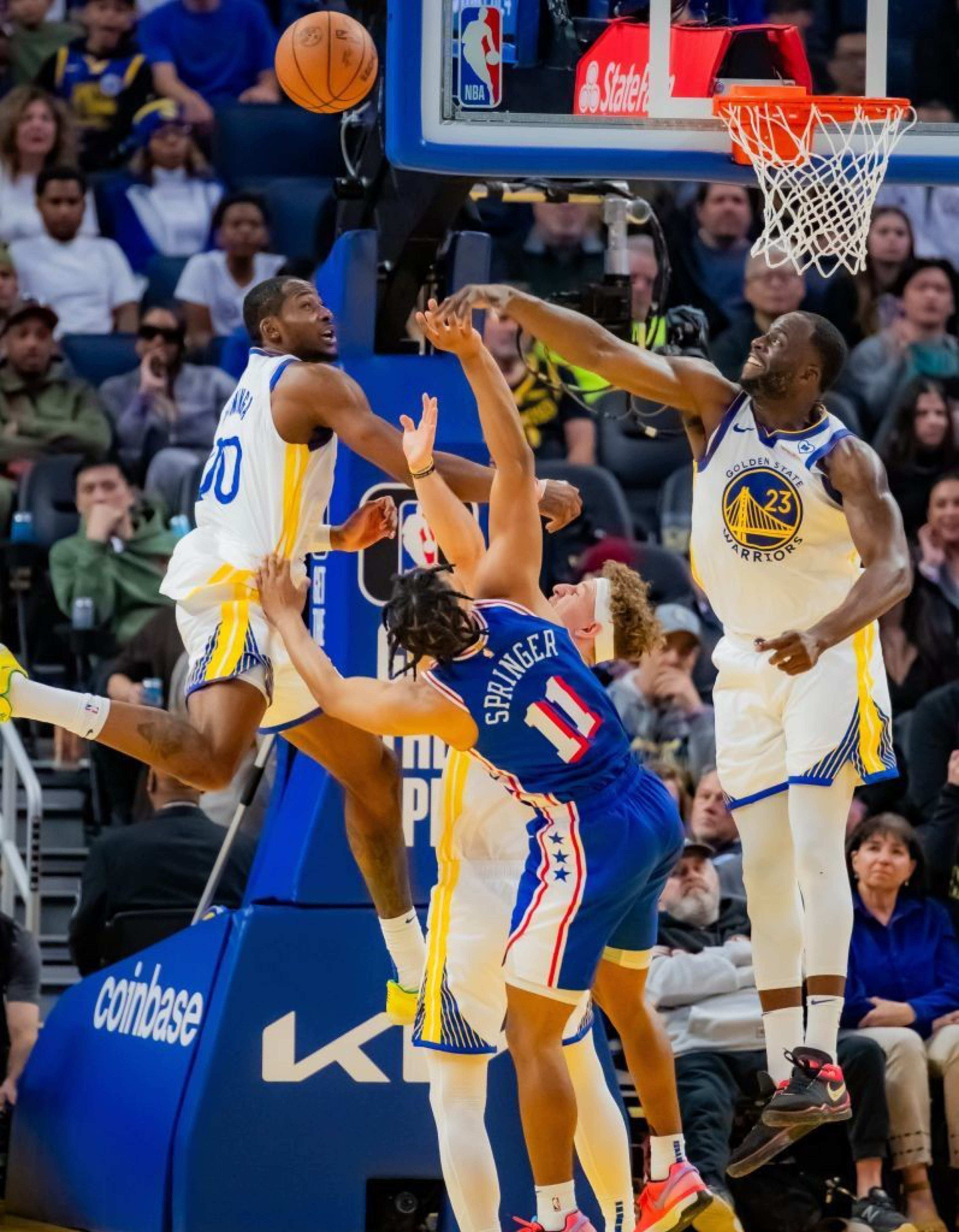 Basketball players from two teams, one in blue and another in white, compete near the hoop. The player in blue, wearing number 11, is being blocked by two players in white.