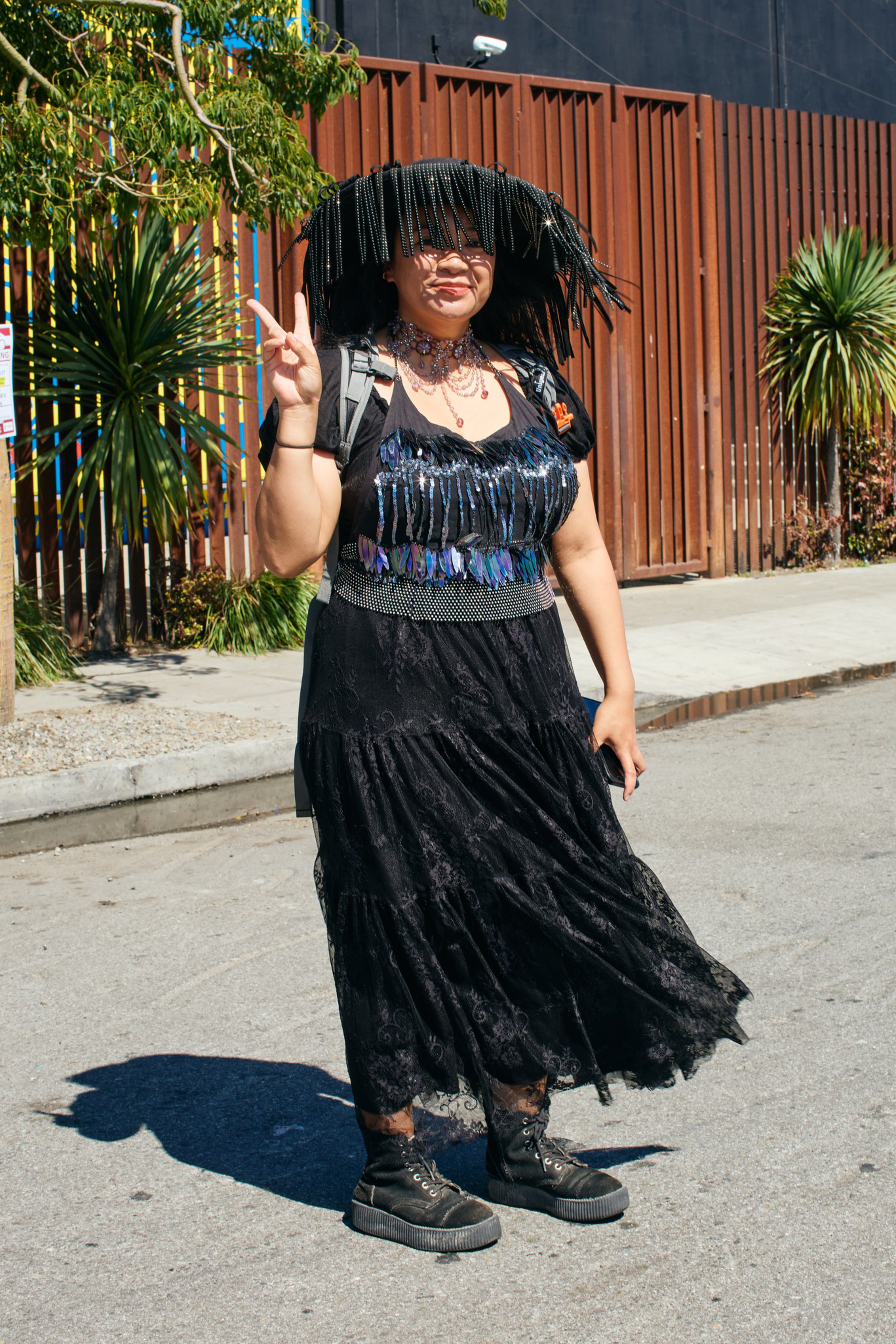 A person wearing a black fringe hat, a black dress with sequins, black boots, and making a peace sign while standing on the street.