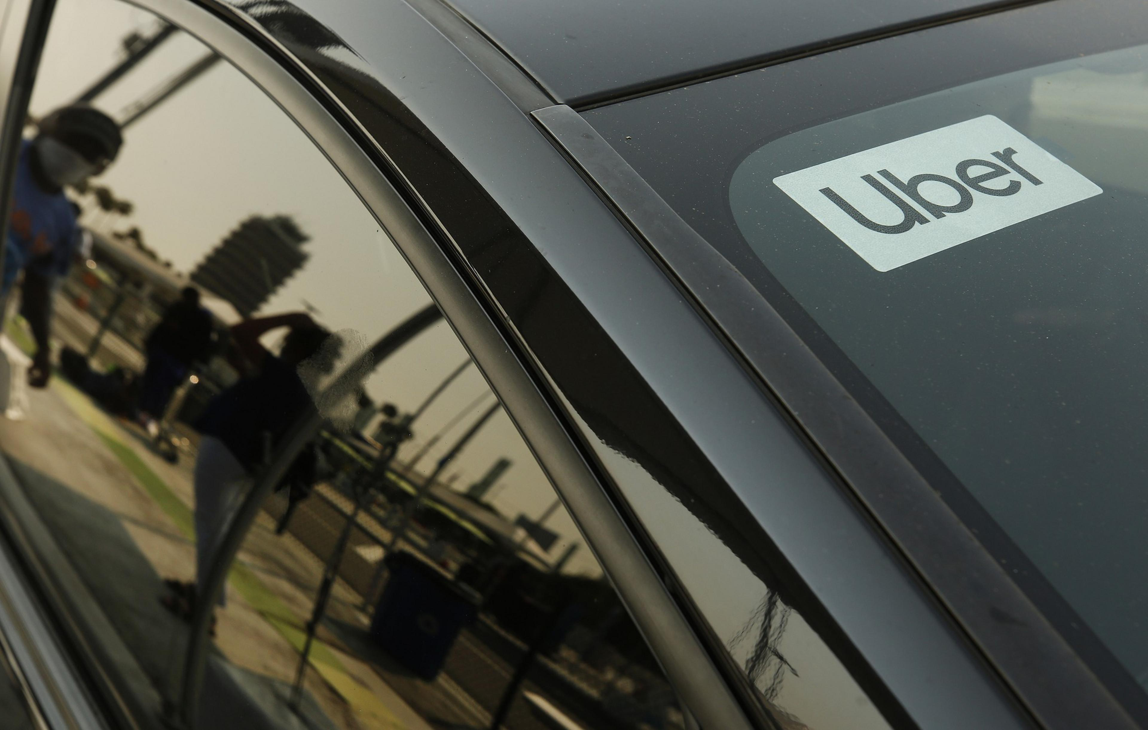 A black car displays an Uber sign in the window. The reflection on the car shows blurred people and a building under a sunny sky.