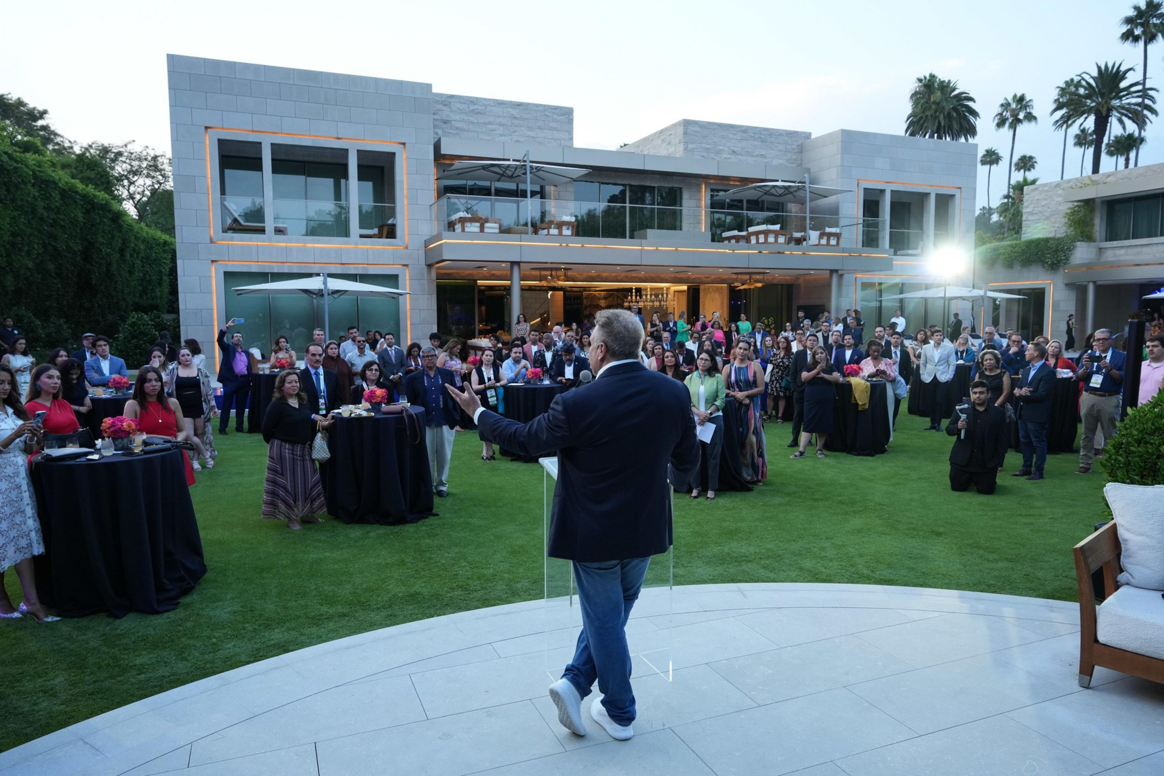 A man speaks to a large audience gathered on a lawn outside a modern, multi-story building during an outdoor event.
