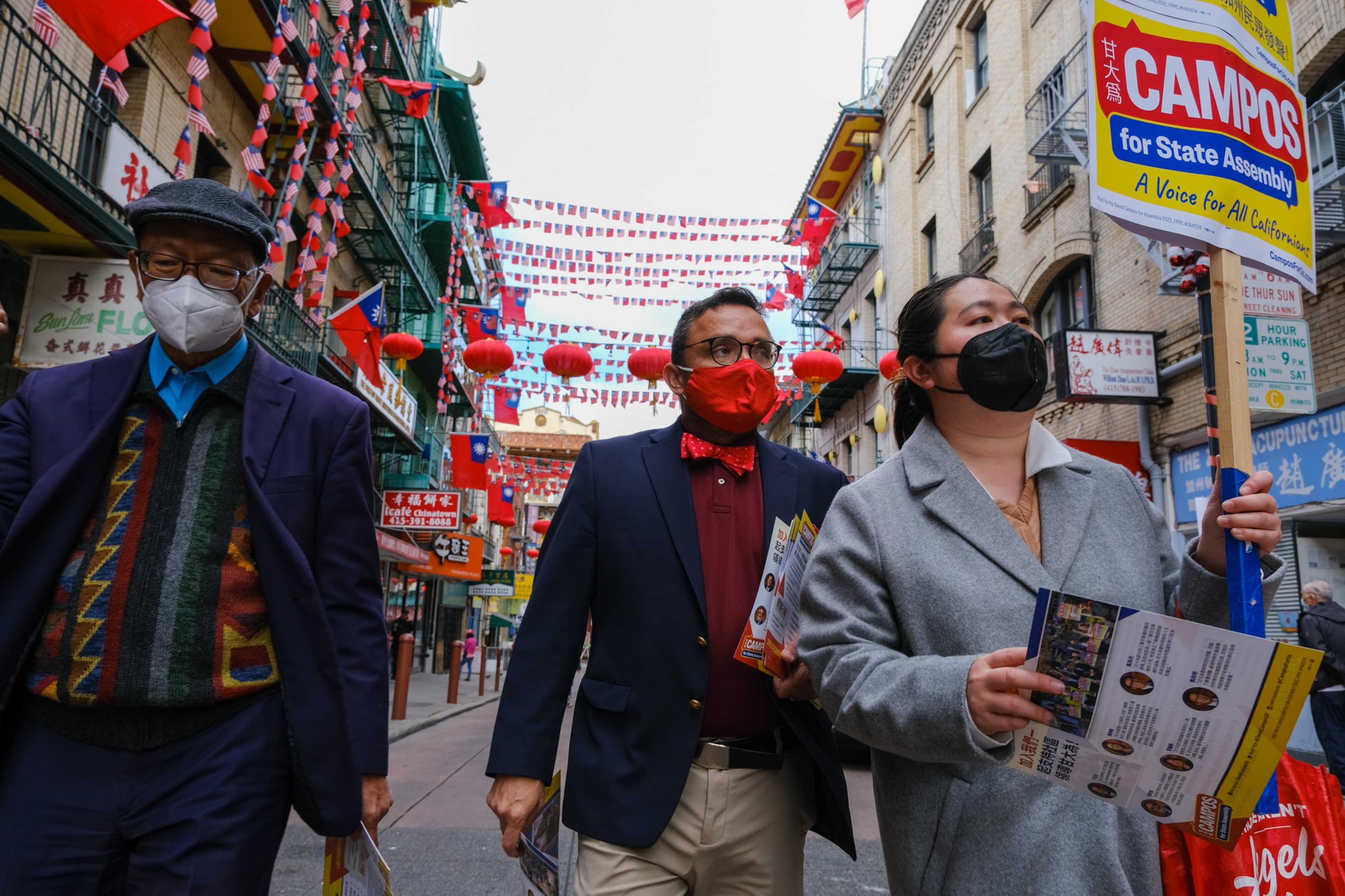 Three people wearing face masks walk down a decorated street with red lanterns and flags, one holding a "Campos for State Assembly" sign and flyers.