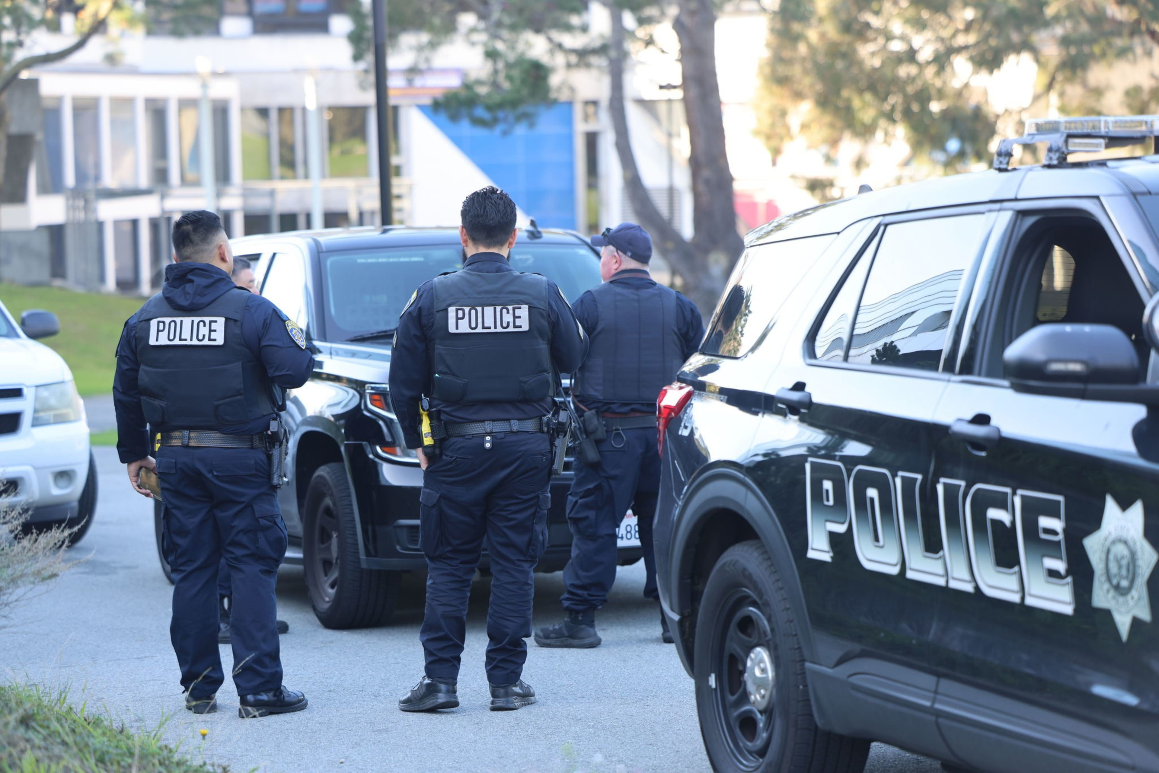 Three police officers stand near police vehicles on a street, wearing uniforms and gear with &quot;POLICE&quot; visible on their vests and cars.
