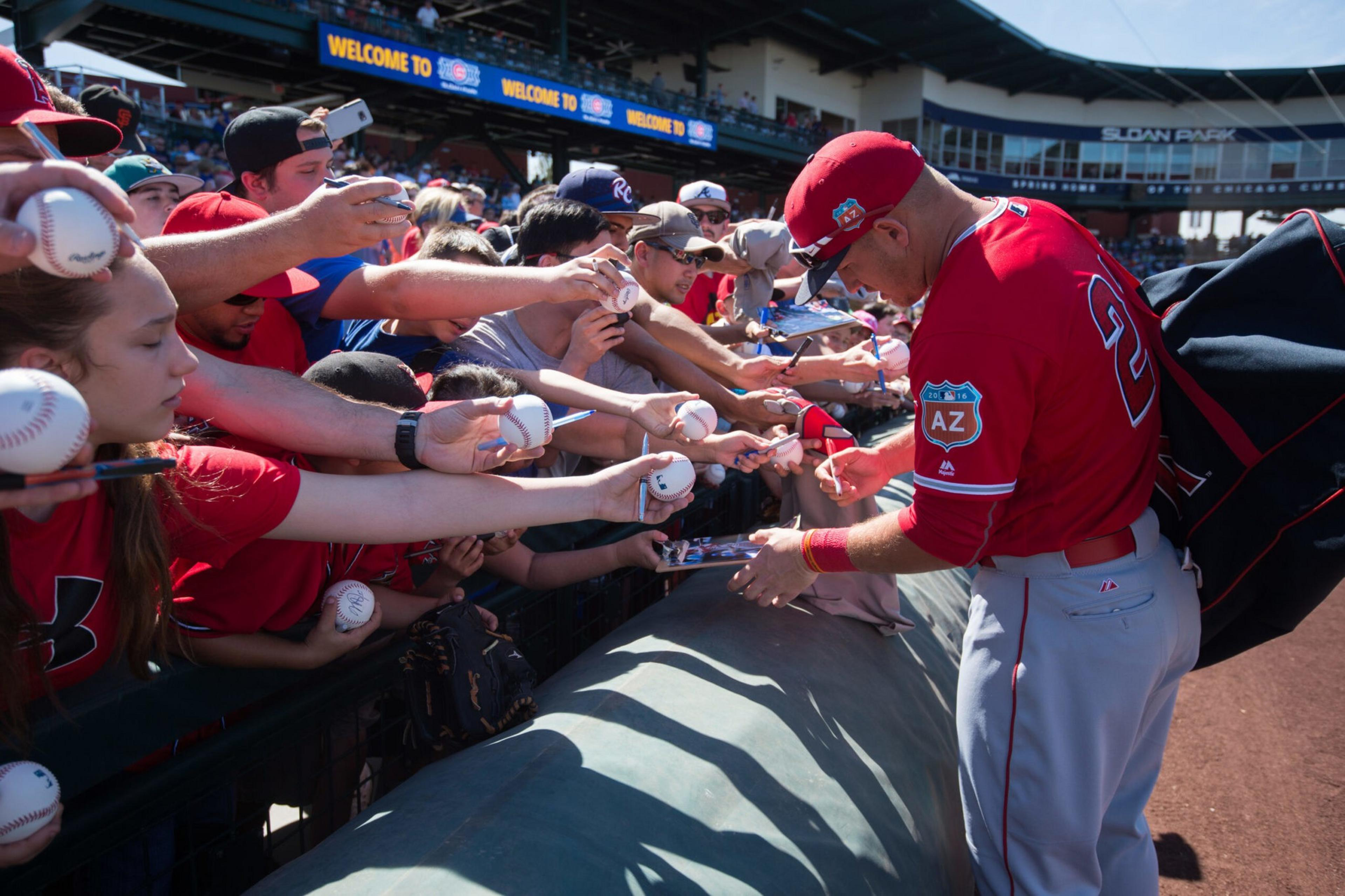 A baseball player in a red jersey signs autographs on baseballs and photos for fans crowded behind a fence at a stadium.