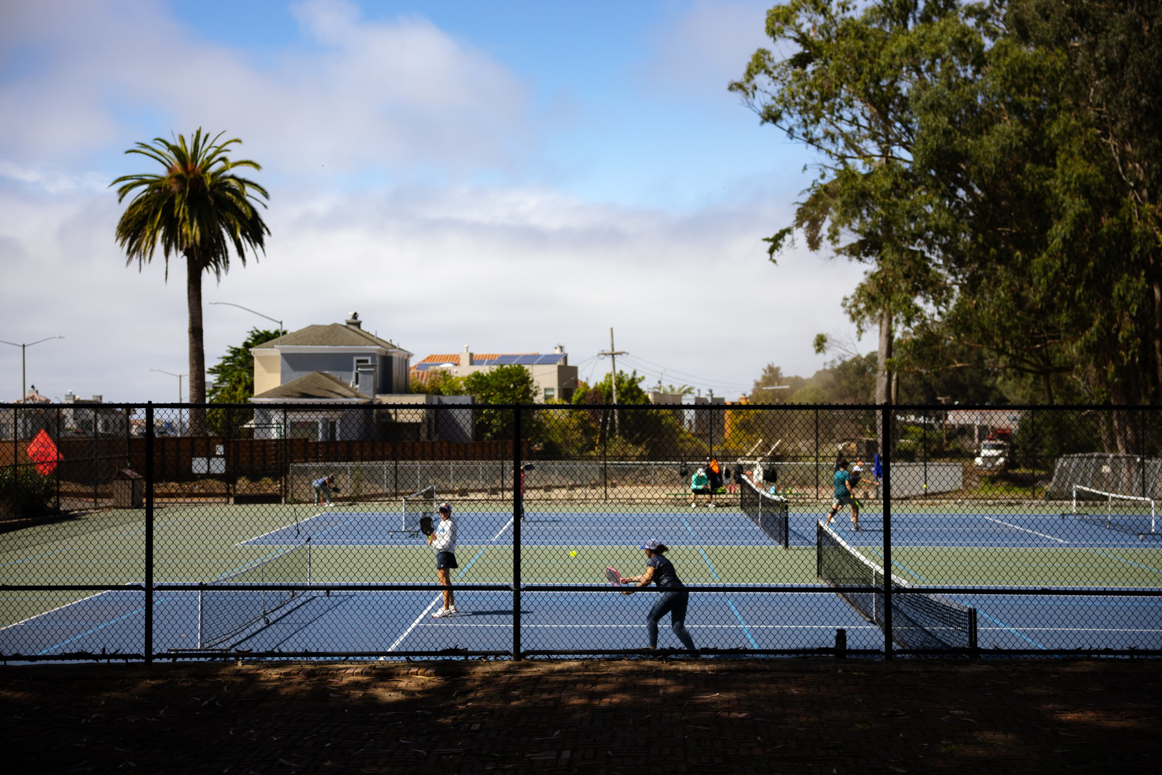 Two pairs of people are playing tennis on adjacent outdoor courts surrounded by a black chain-link fence, with trees and houses in the background.