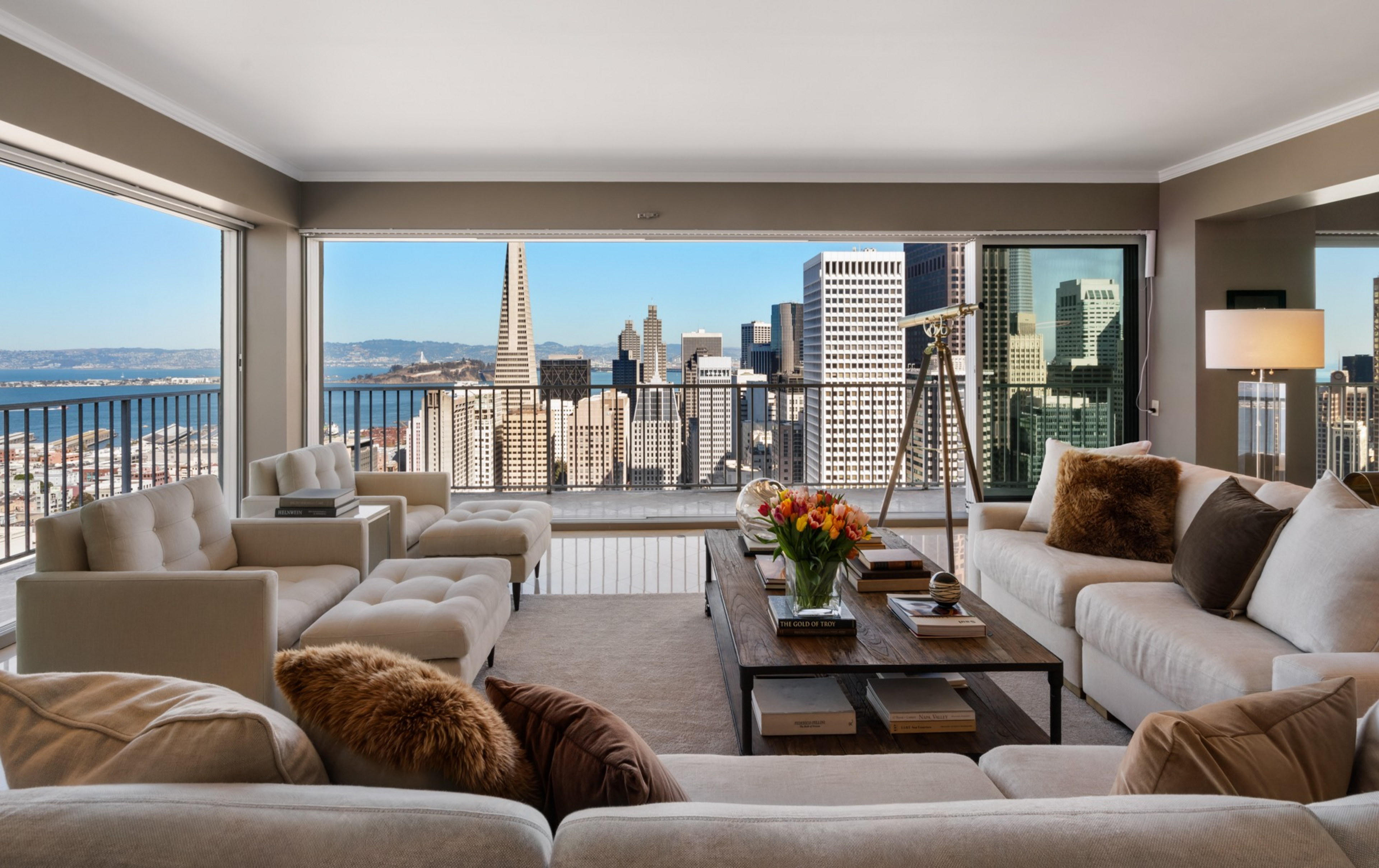 A modern living room with beige sofas, a wooden coffee table with flowers, books, and a telescope, overlooking a city skyline and water through large windows.