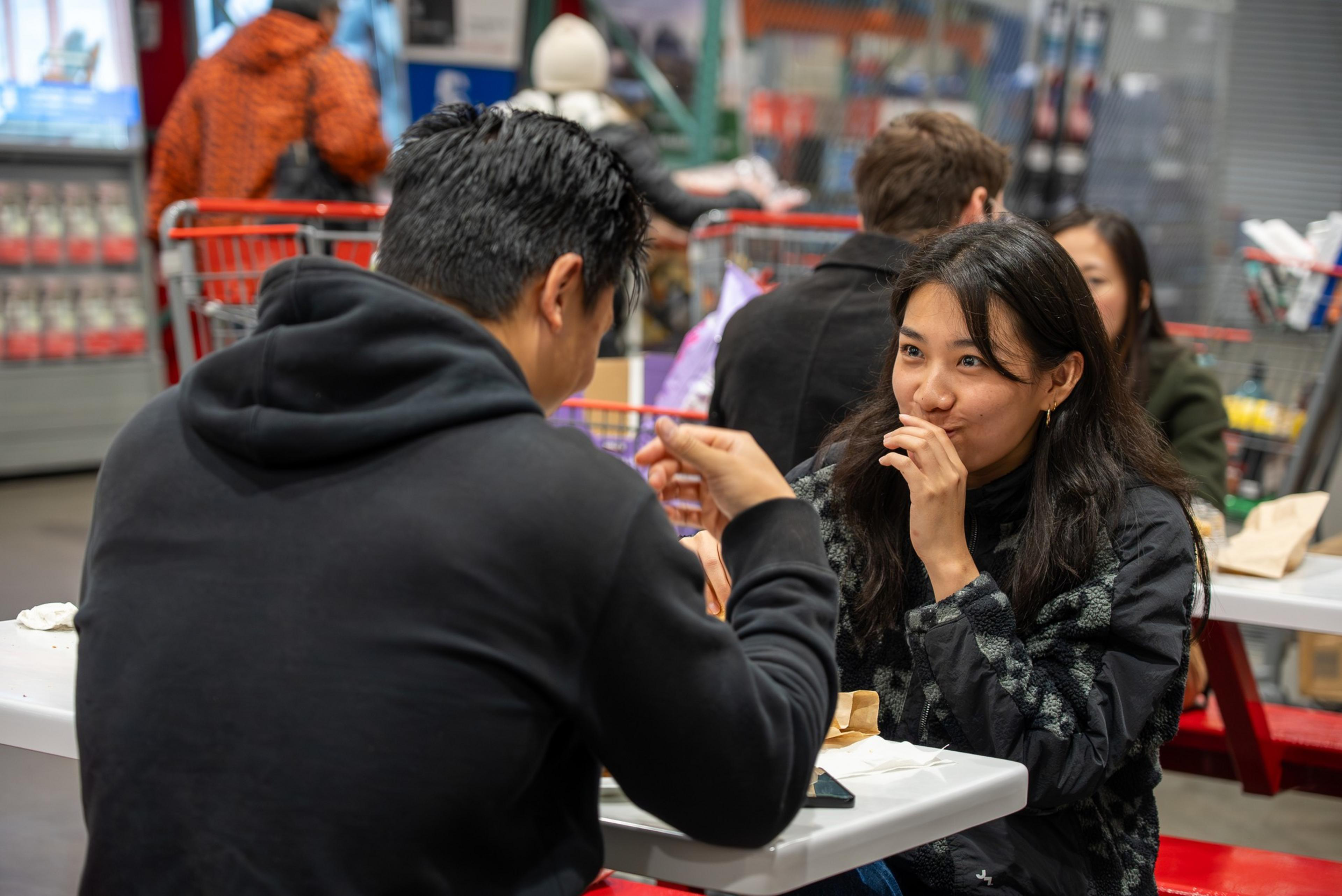 Two people in black jackets sit at a table in a busy indoor market, sharing a meal and smiling while engaged in conversation.