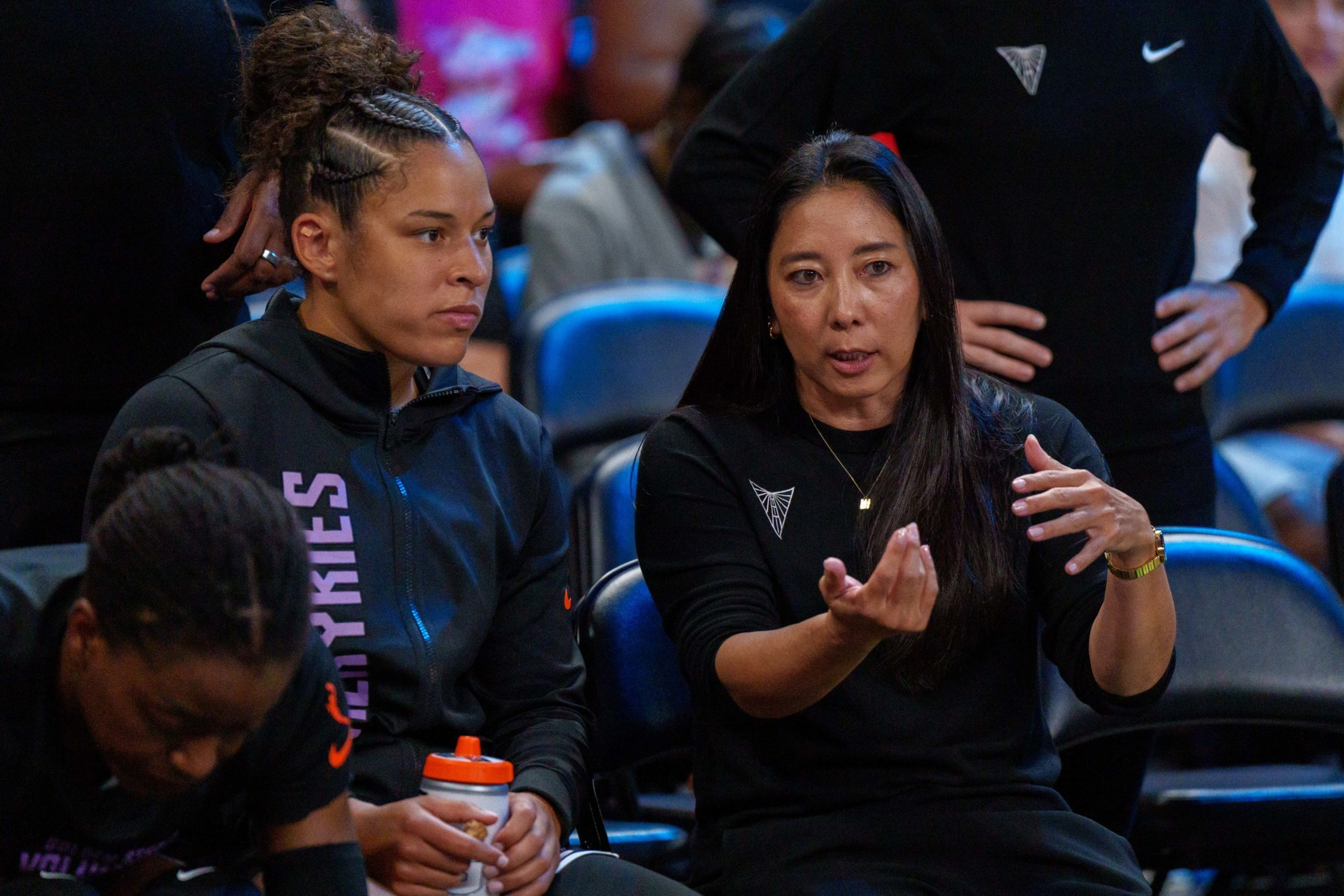 Two women in black sportswear sit talking seriously, one gesturing with her hands, in a setting with blue chairs and other people nearby.