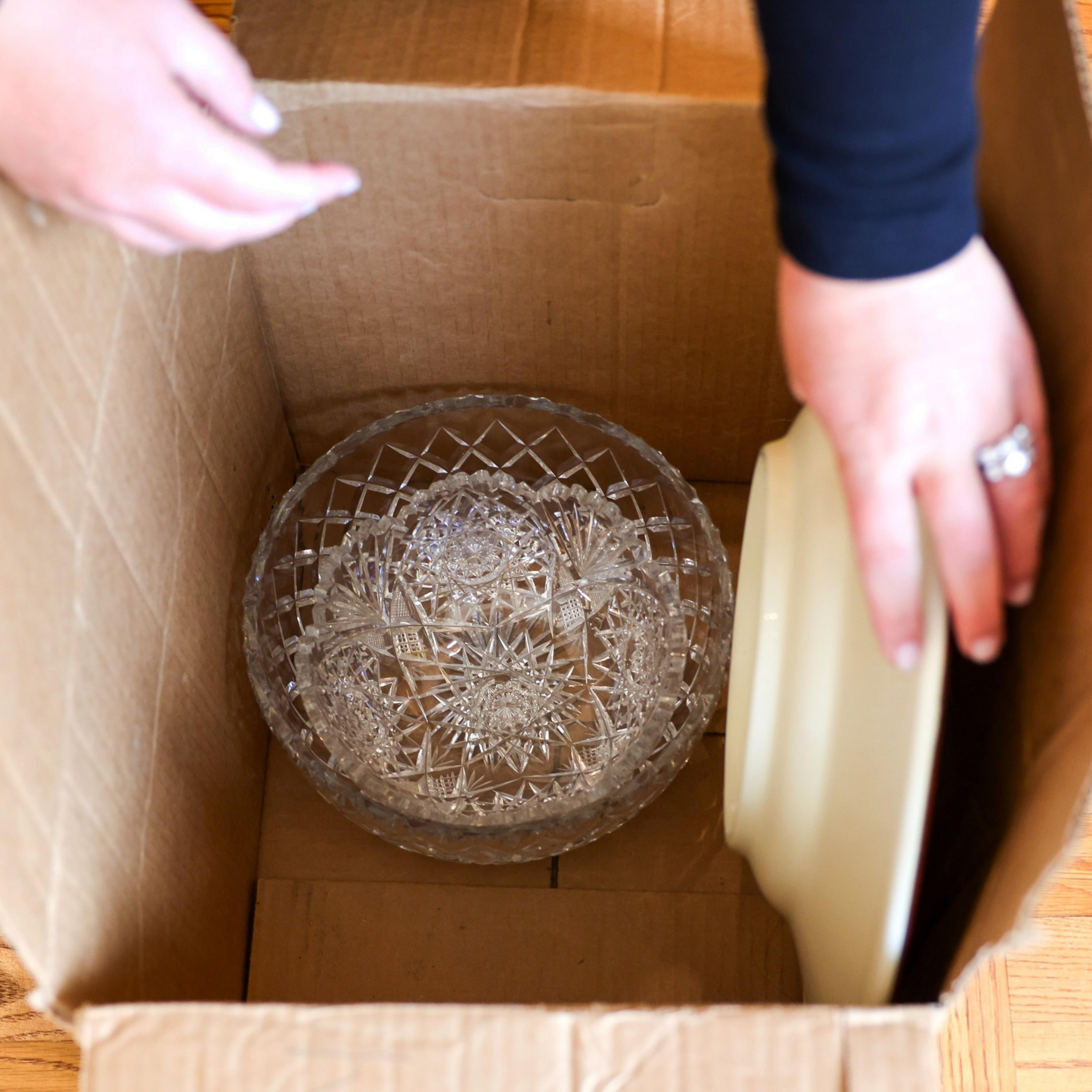 A person's hands holding a beige plate are placing it into a cardboard box containing an intricate, clear glass dish with a decorative pattern.