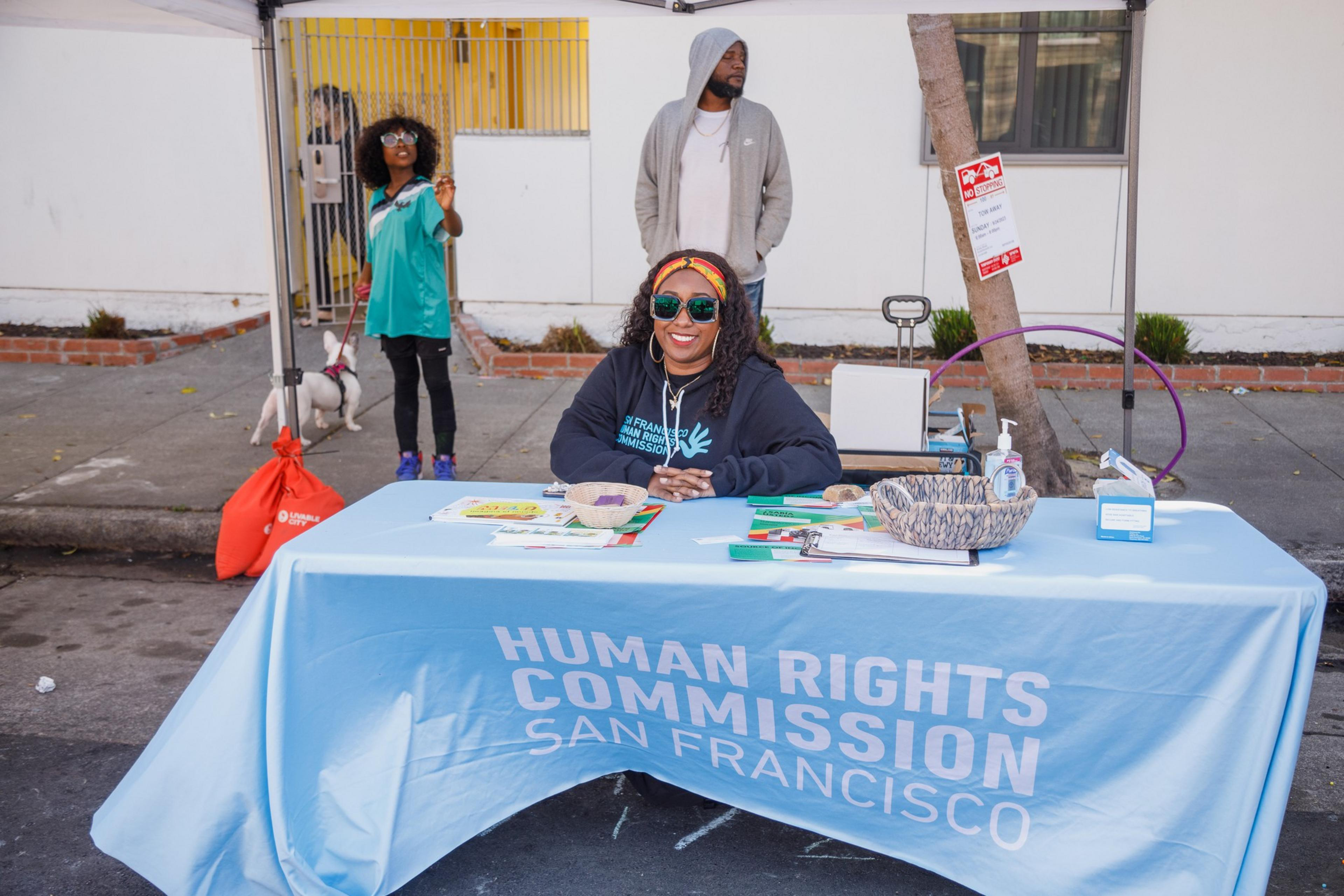 A person sits at a table with a banner &quot;Human Rights Commission San Francisco.&quot; Others stand in the background.