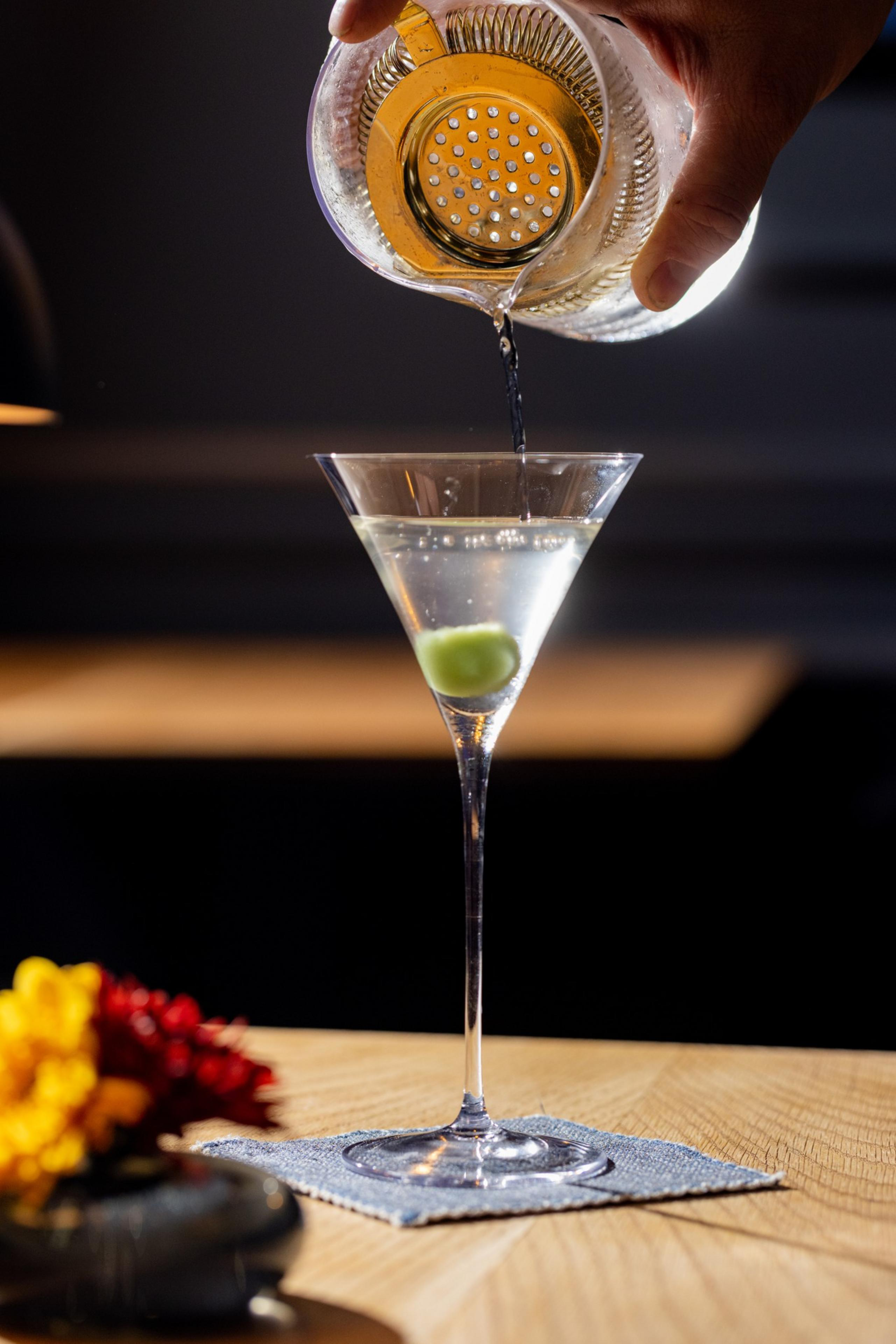A hand pours a cocktail from a shaker into a martini glass with an olive. The glass sits on a table with a small bouquet of red and yellow flowers nearby.