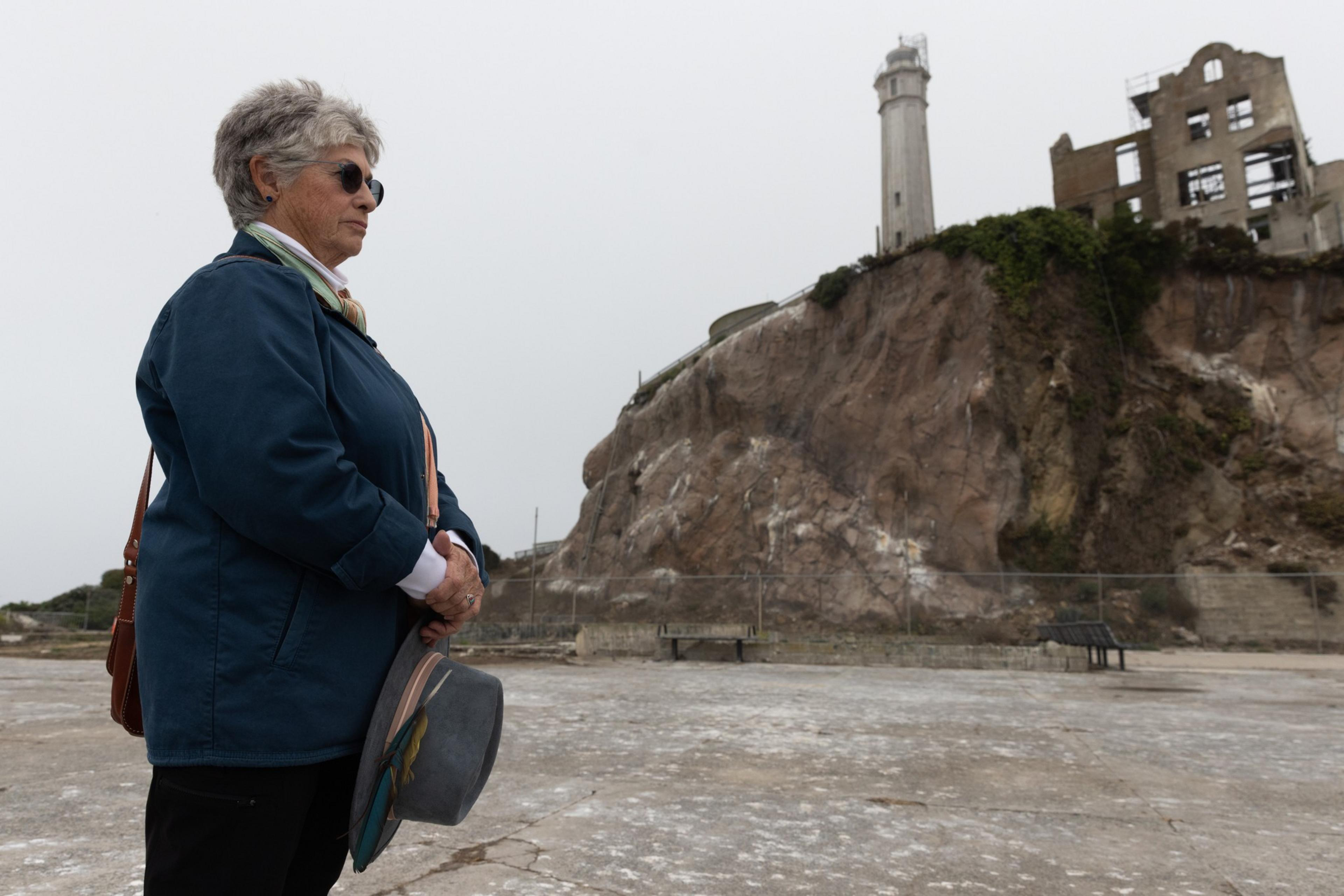 An older woman in sunglasses and a blue jacket stands holding a hat, facing a rocky hill with a tall lighthouse and a partially ruined building.