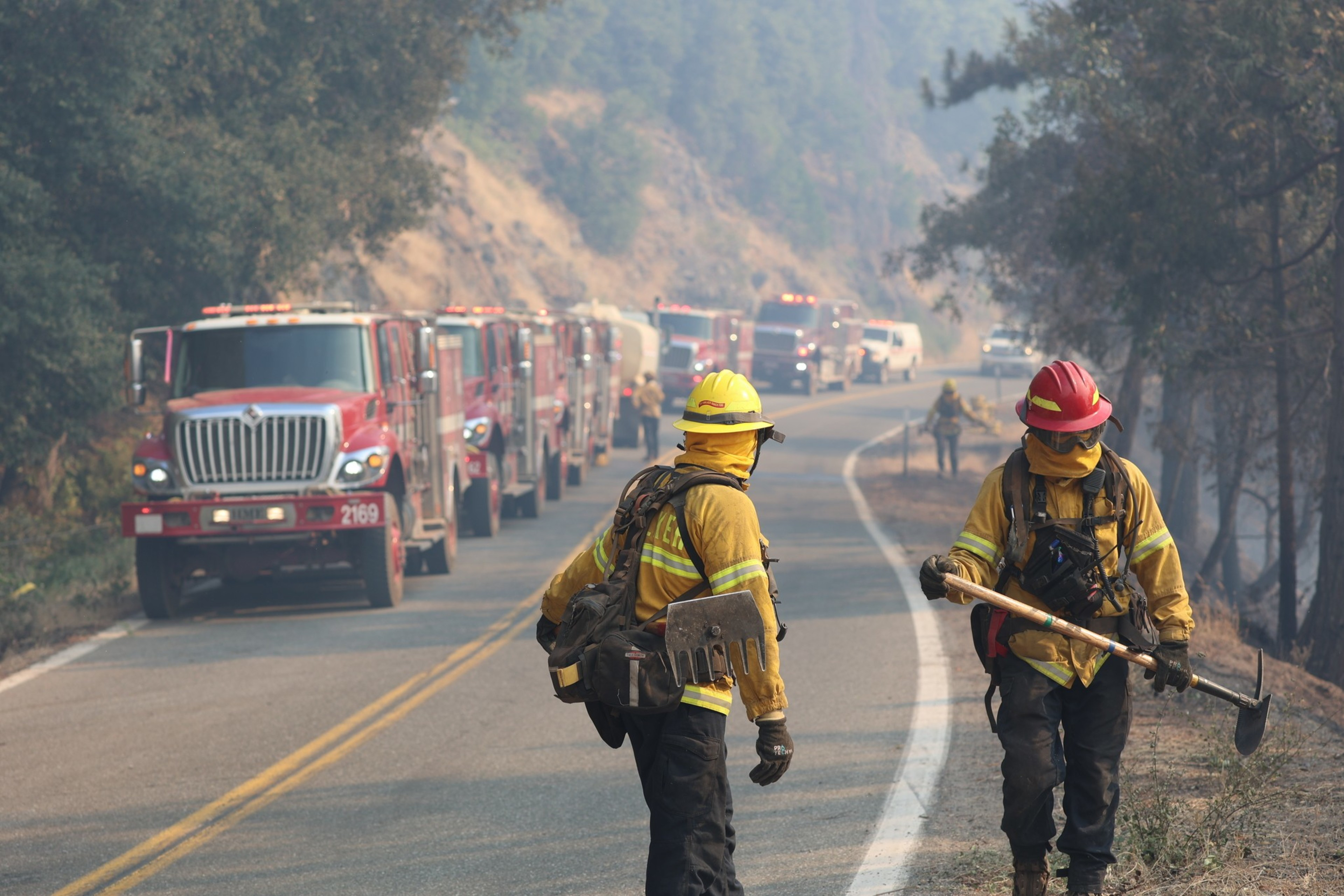 Firefighters stand along a road and several firetrucks are lined up near them.