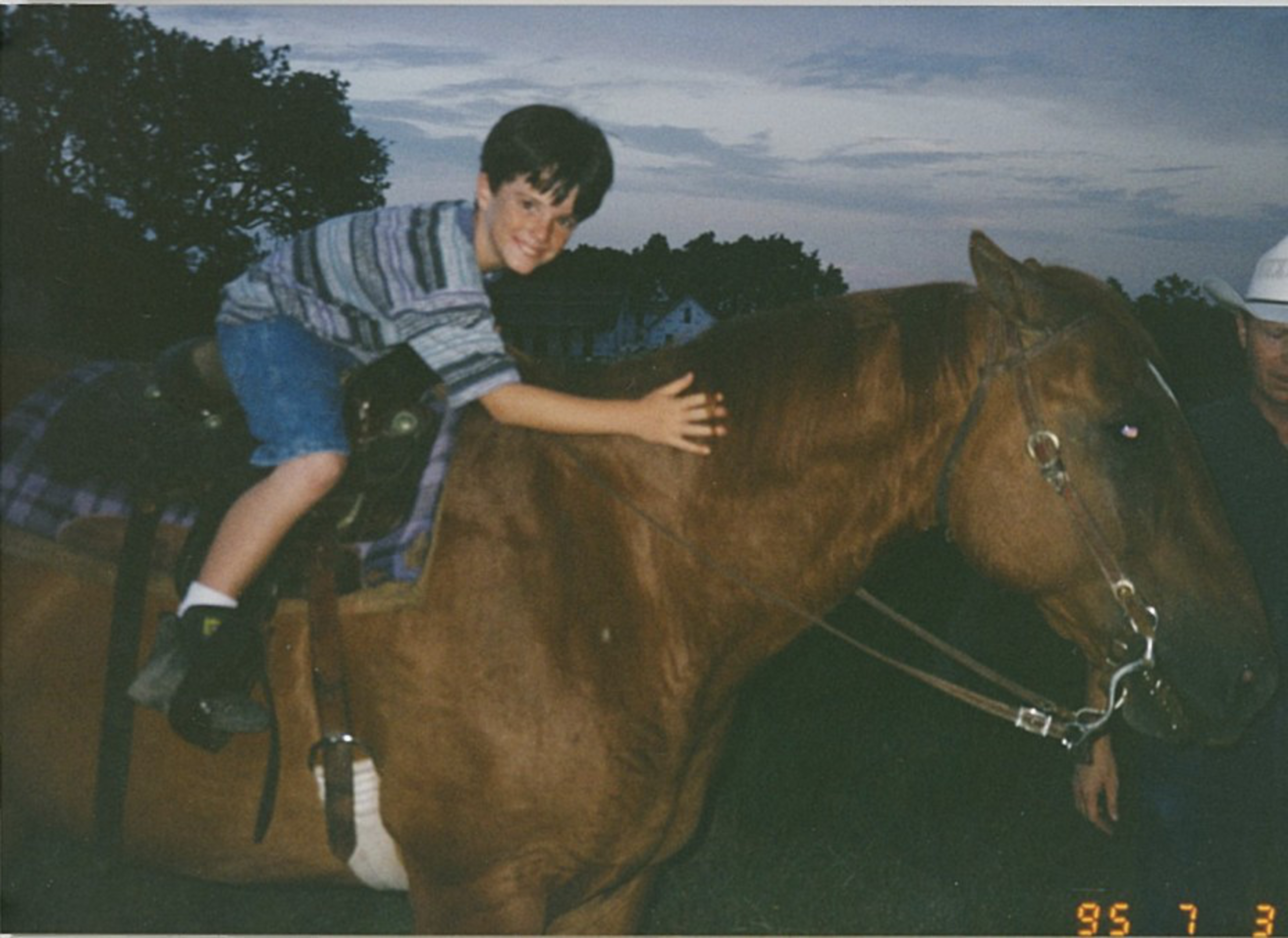 A young boy, wearing a striped shirt and blue shorts, is happily sitting on a brown horse during dusk, while a man in a white hat stands beside them.