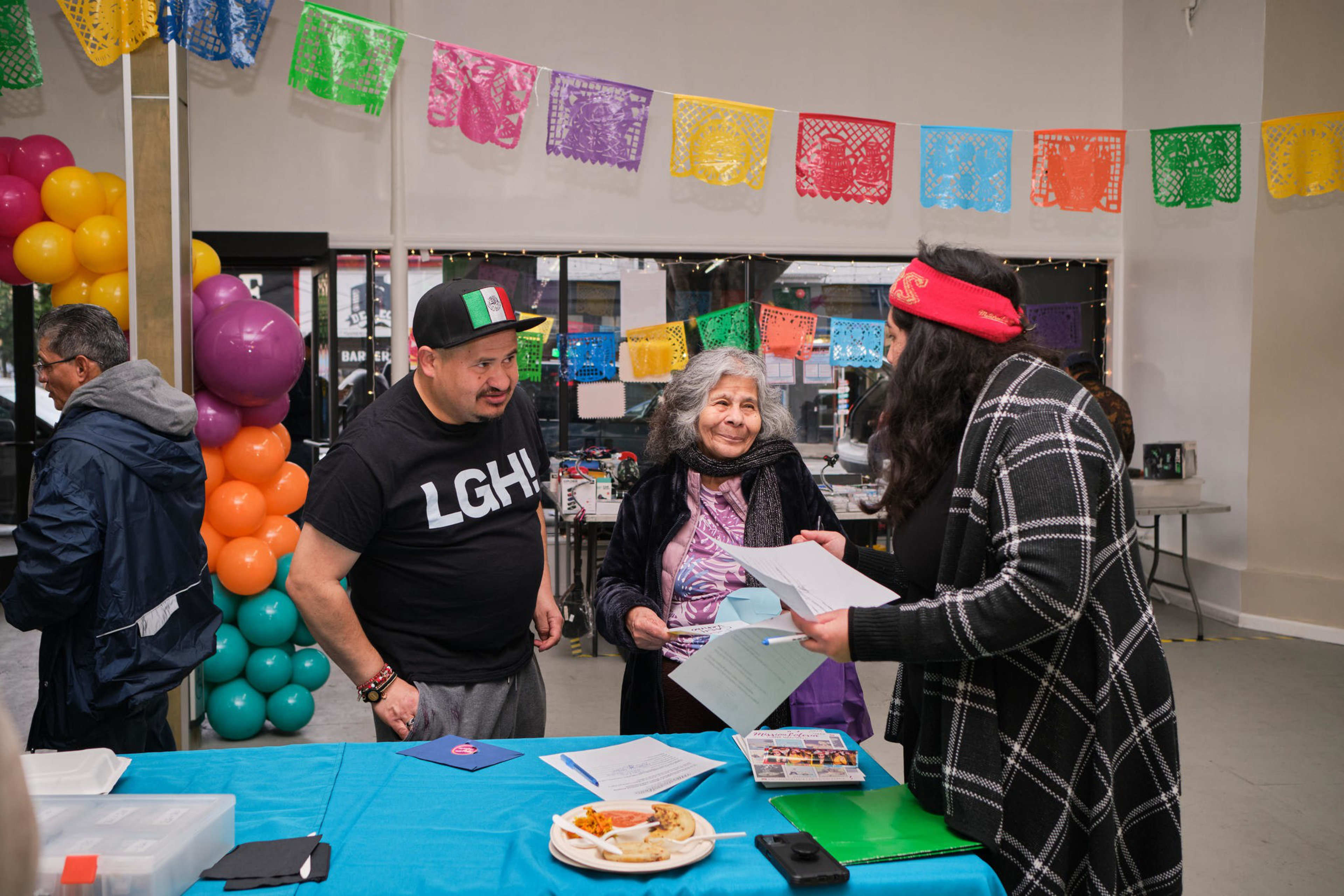 Three people converse near a table with colorful decorations, balloons, and papers at an indoor event.