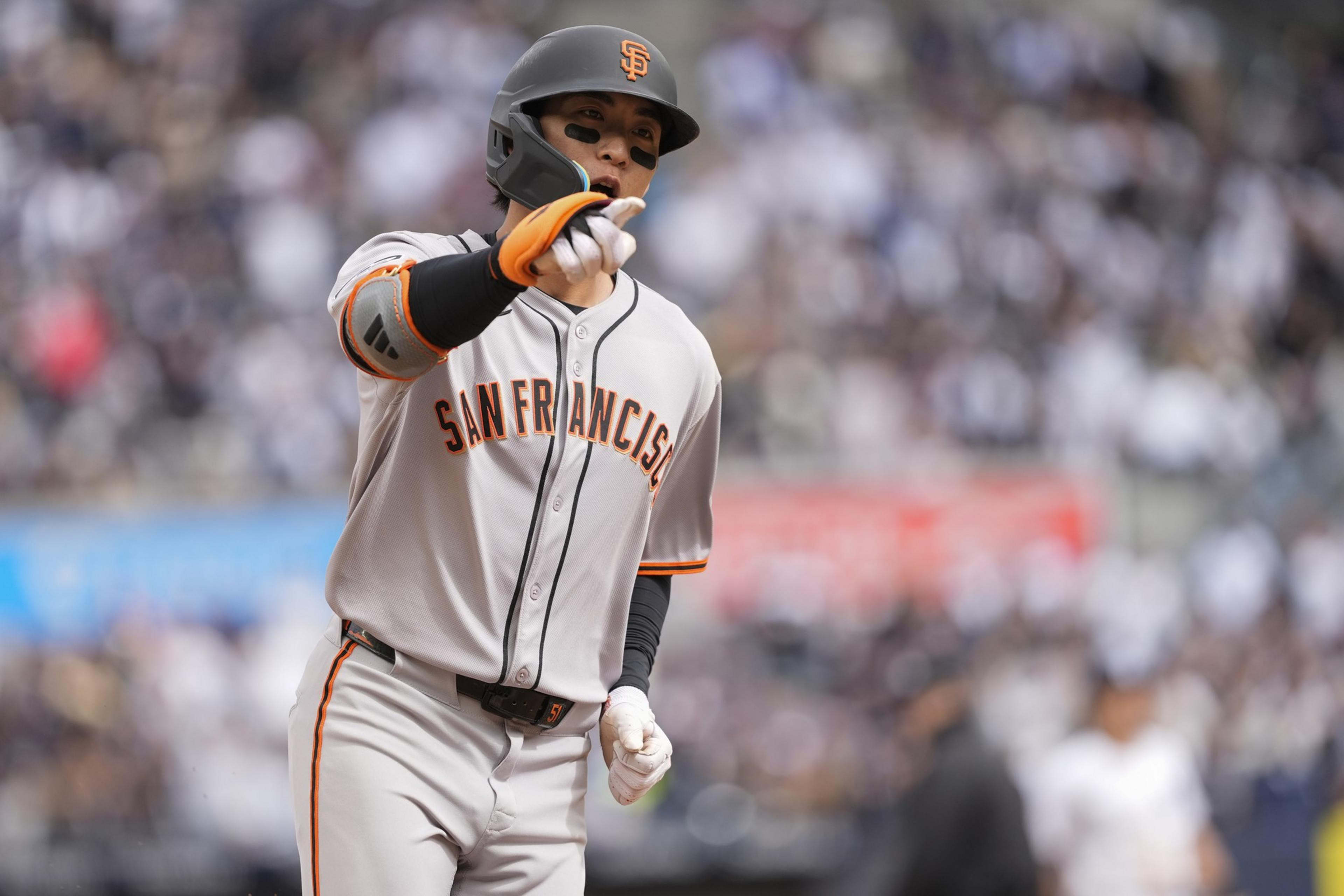 A baseball player in a San Francisco uniform, wearing a helmet and black face paint, points forward. The background is blurred with a crowd.