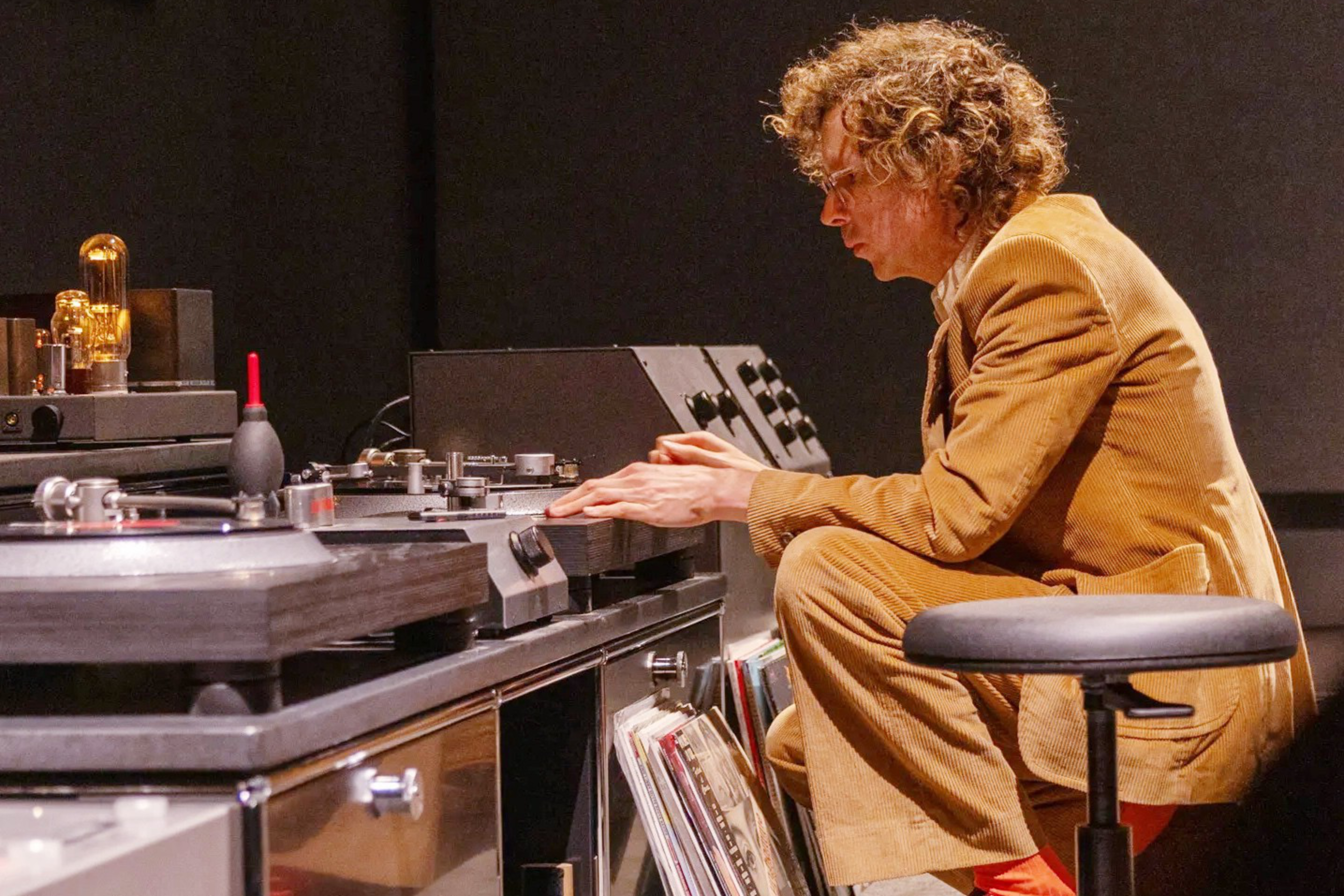 A person with curly hair, wearing a brown suit, is adjusting equipment on a modern sound mixing desk. Vinyl records are stored nearby, and a stool is in the foreground.