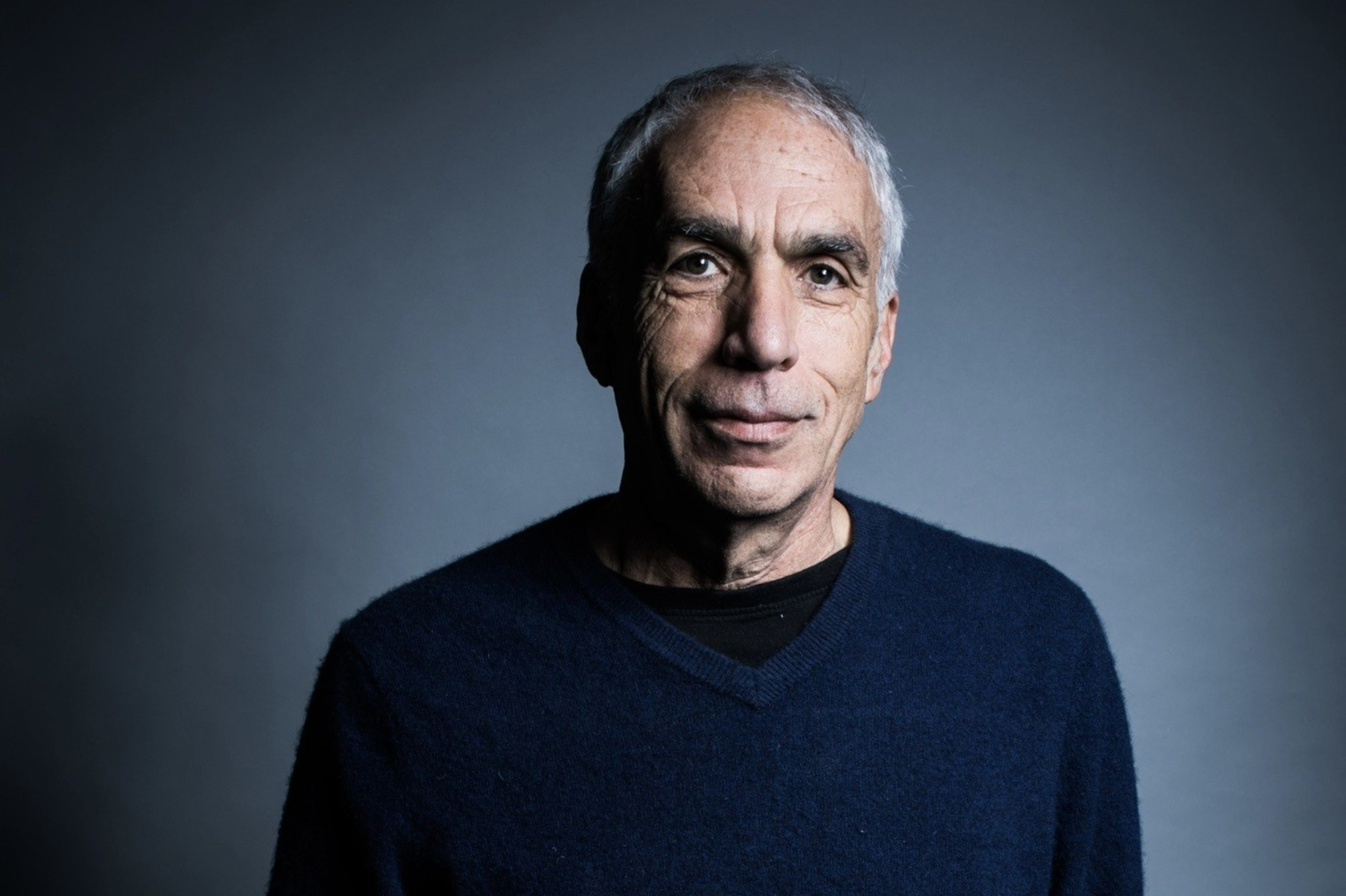 An older man with short gray hair is wearing a dark blue sweater and jeans, standing with hands in pockets against a plain gray background.