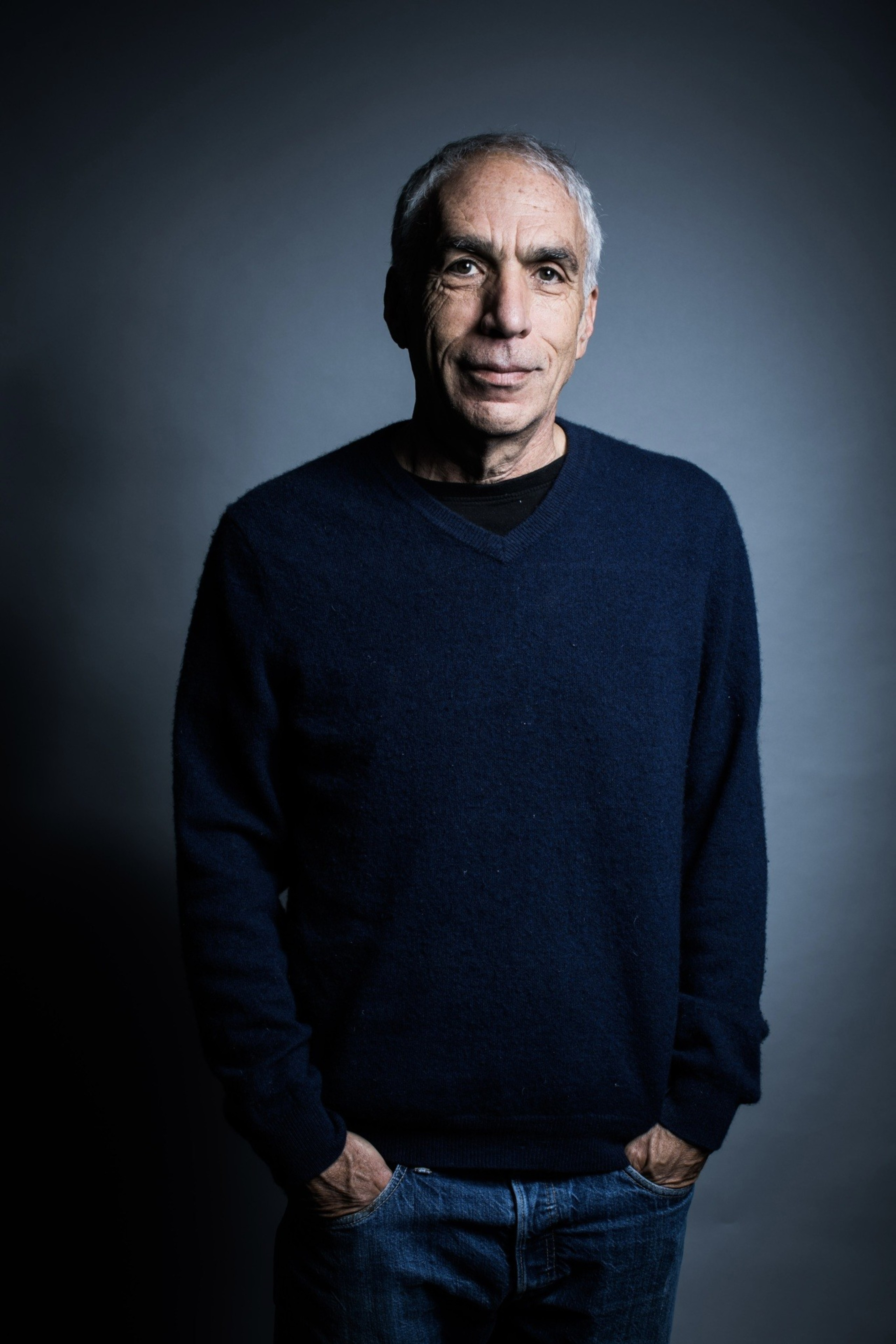 An older man with short gray hair is wearing a dark blue sweater and jeans, standing with hands in pockets against a plain gray background.